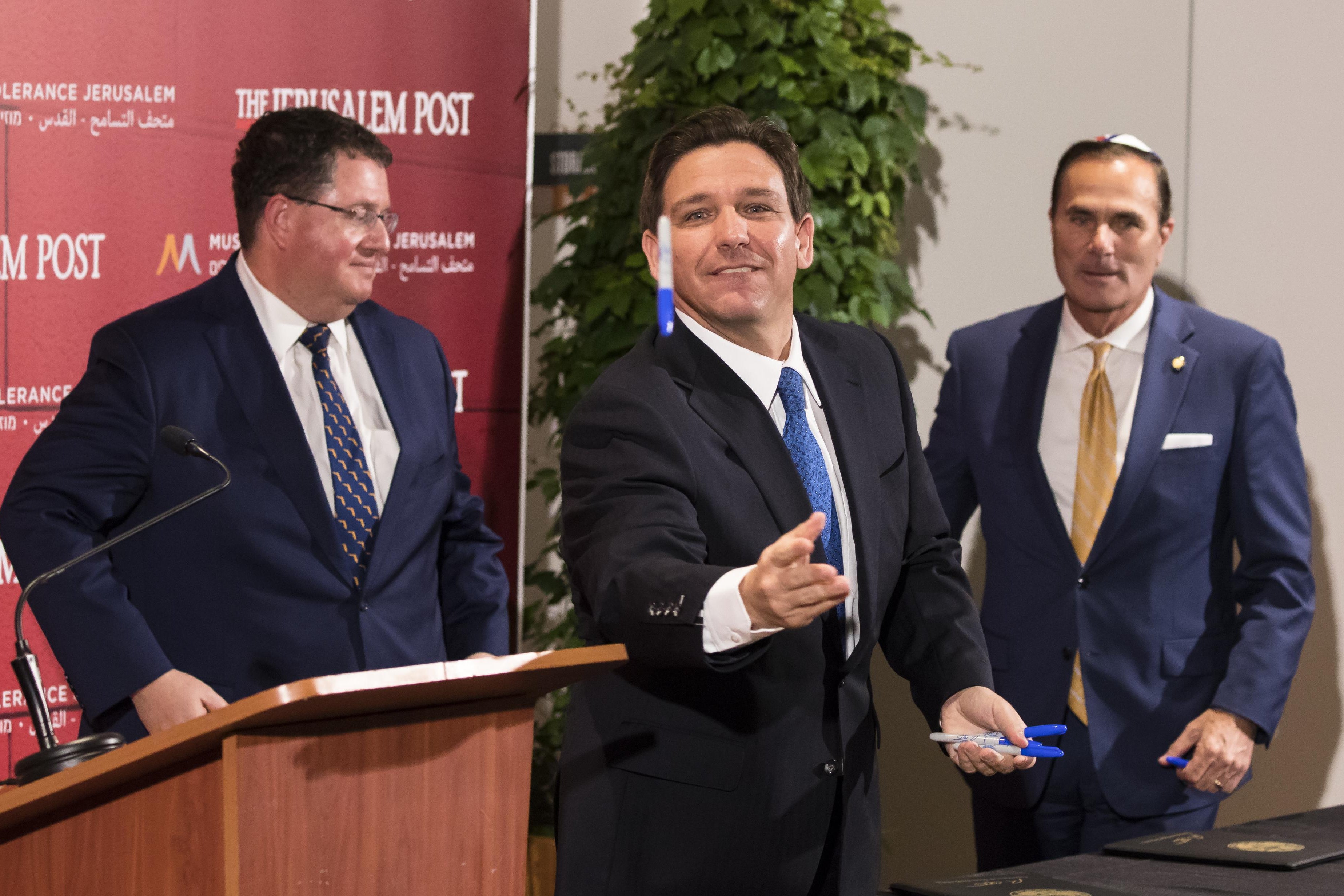 Ron DeSantis stands next to a podium and throws a blue Sharpie with one hand. In his other hand, he holds several more blue Sharpies. Two men stand in the background.