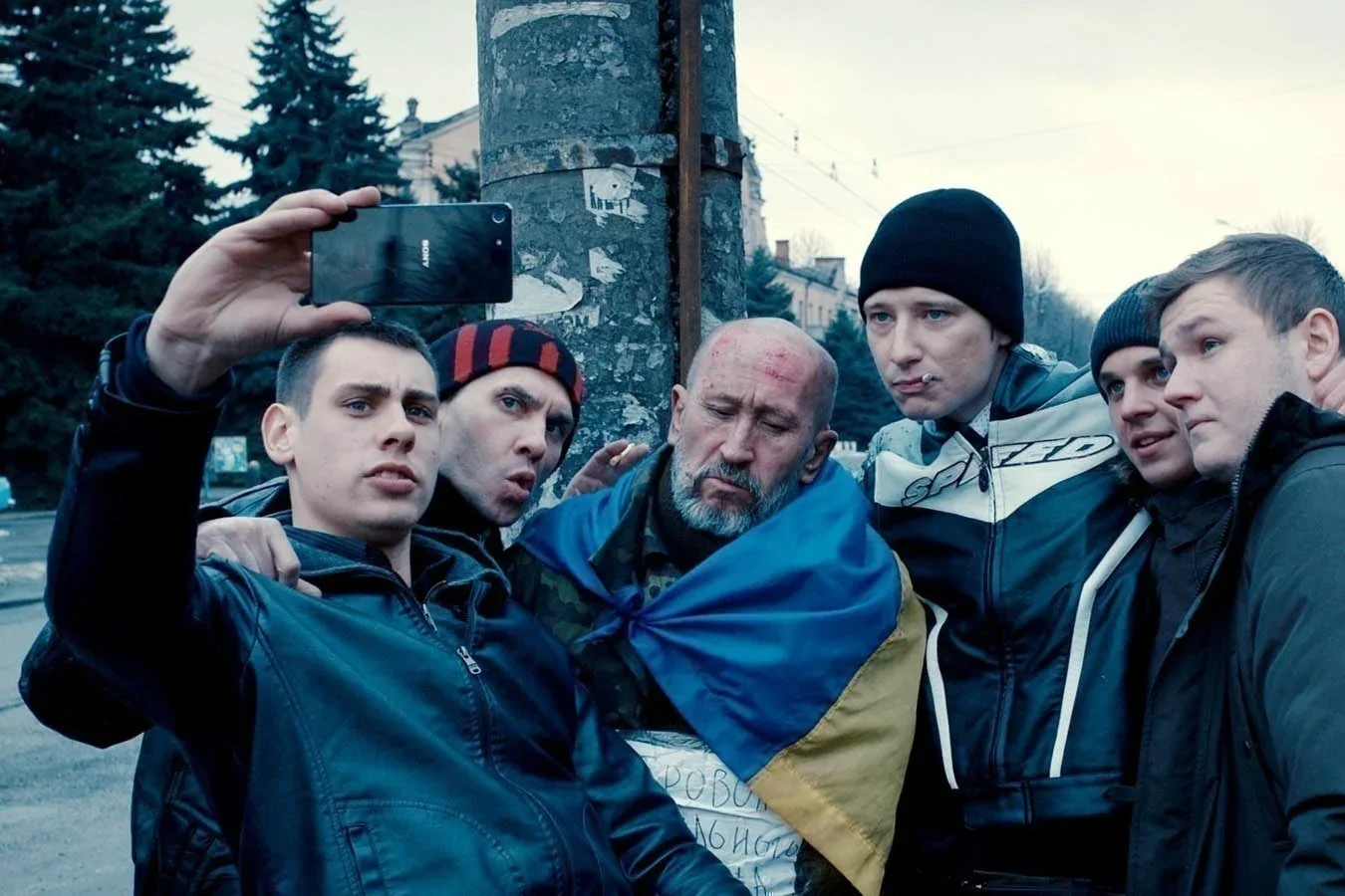 Five relatively young men pose with an older man draped in a Ukrainian flag for a selfie. The man in the Ukrainian flag looks beaten down, as if he's been forced into this.