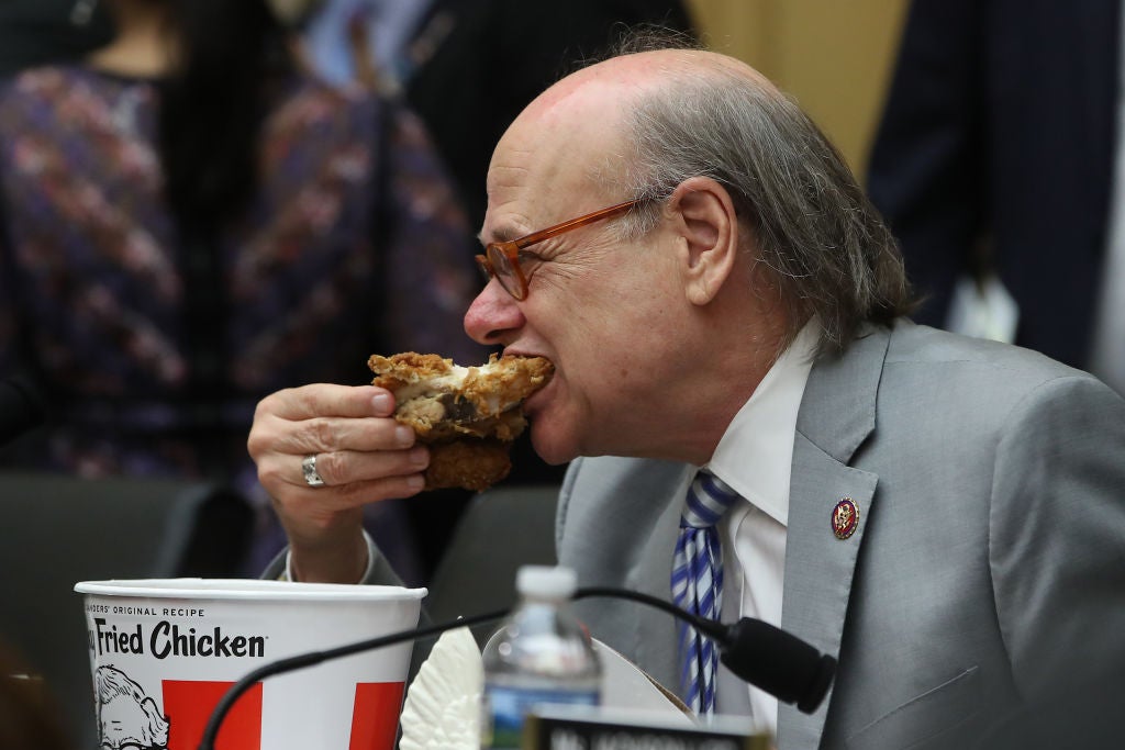 Cohen, a balding gentleman wearing a gray suit, takes a large bite of a KFC chicken breast while seated with a KFC bucket next to him on a dais.