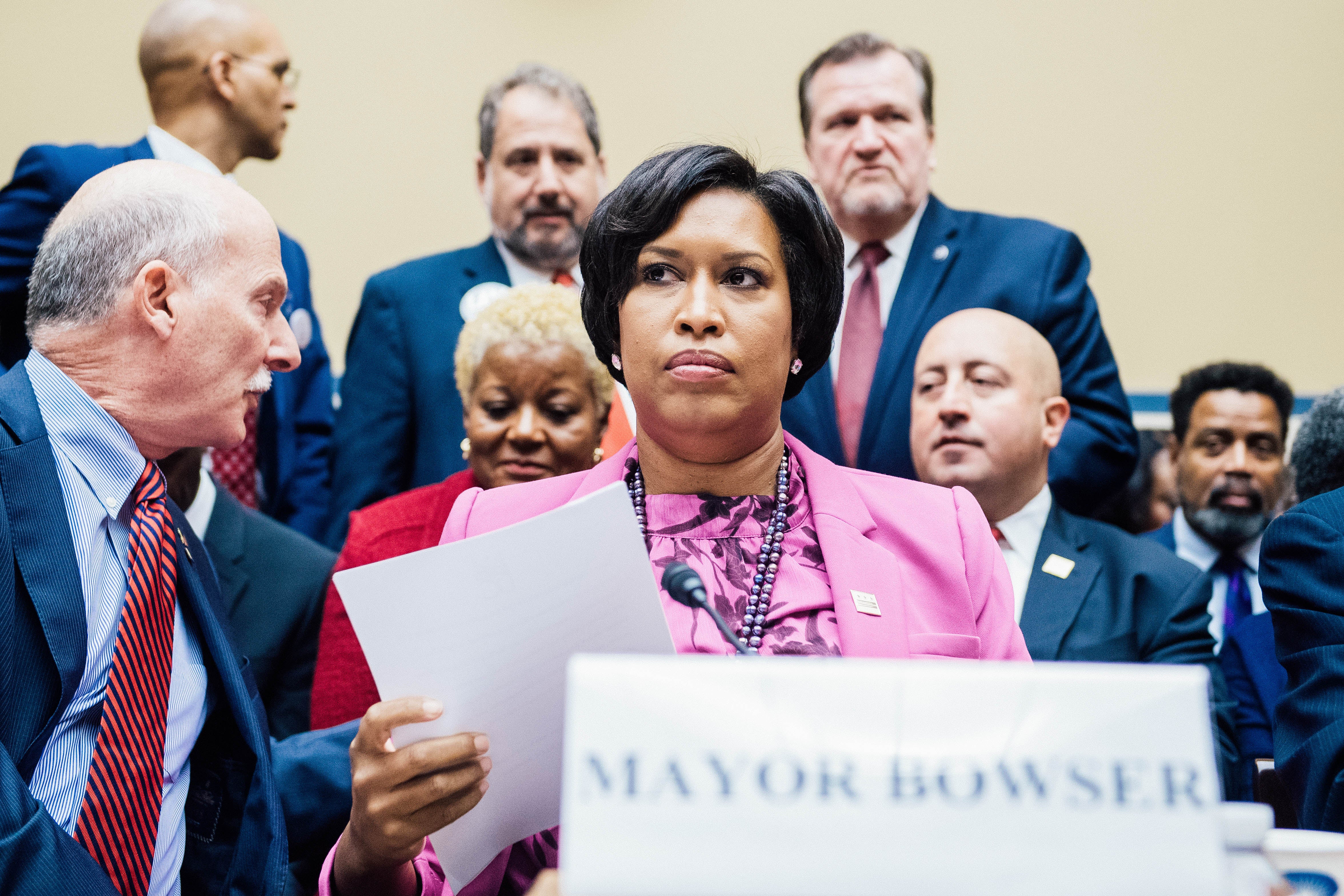 Mendelson turns backward in his seat toward other people at the hearing. Next to him, Bowser holds a piece of paper and sits behind a placard that reads "MAYOR BOWSER."