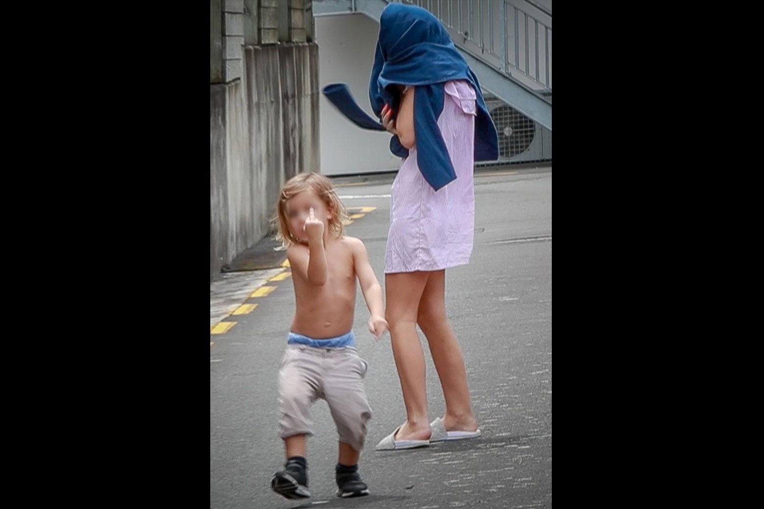 A shirtless young boy flips off the camera. Beside him, a female relative covers her face with pants.