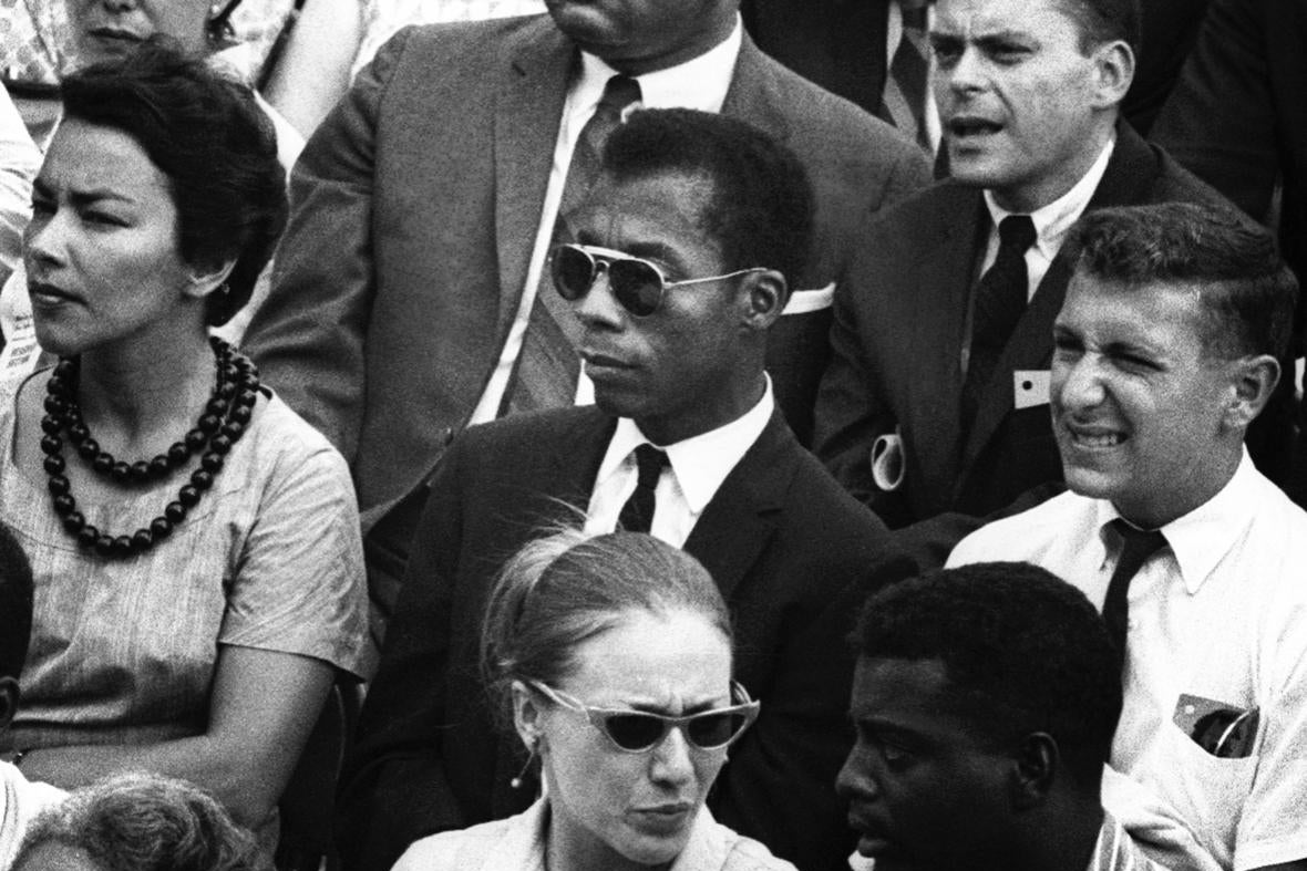 James Baldwin is seated in a crowd, wearing black sunglasses. The image is black and white.