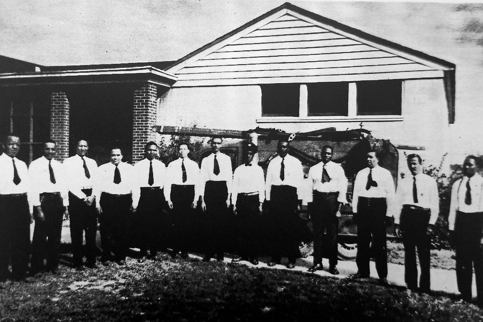 Thirteen men in white shirts and ties stand in a line in front of a low building.