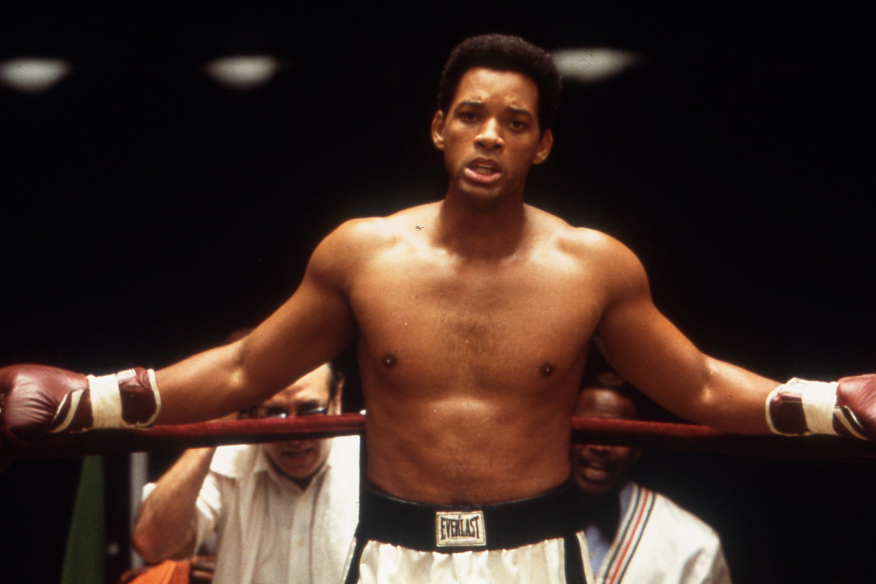 Will Smith leans on the ropes of a boxing ring, wearing boxing gloves and boxing shorts.