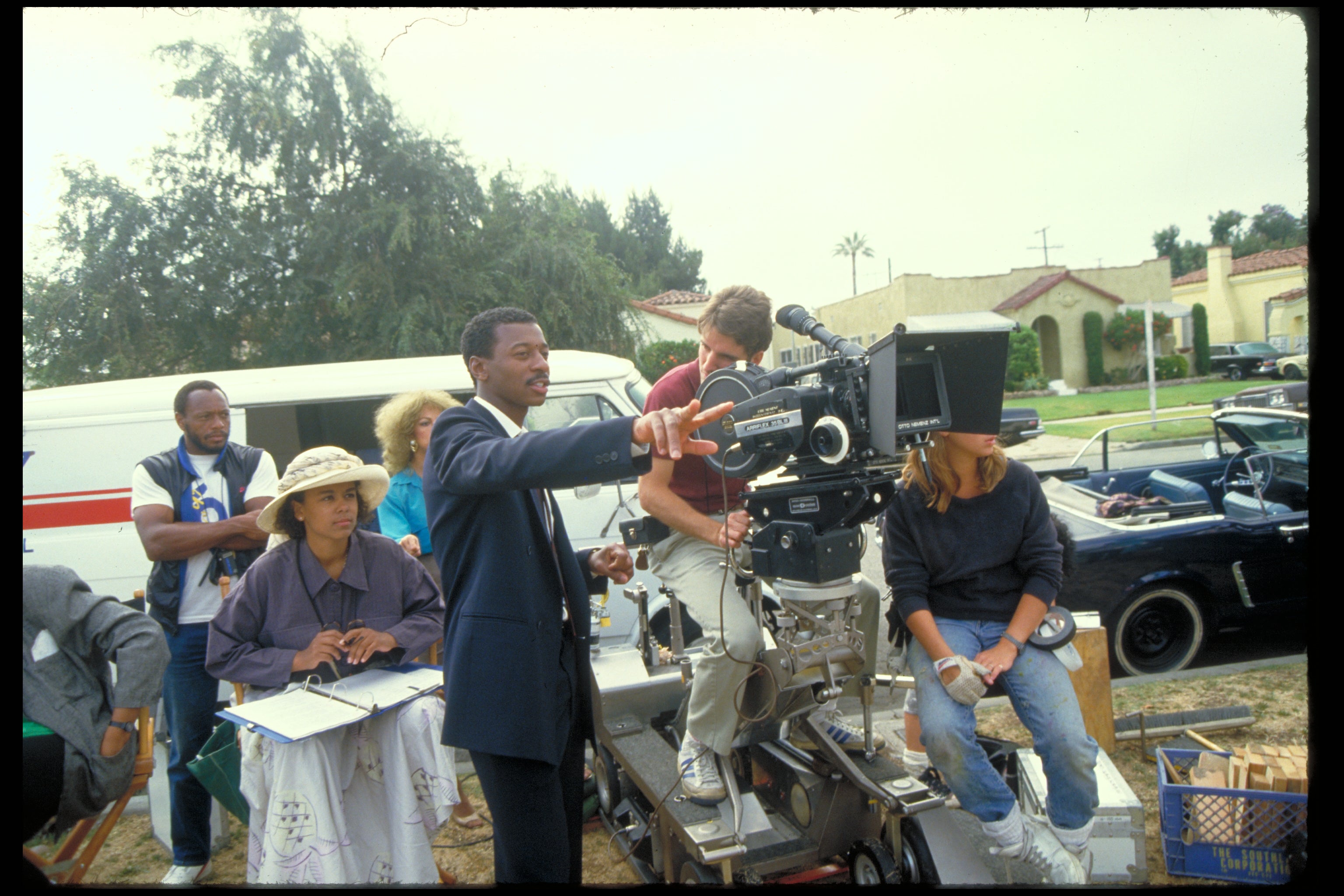 A Black man in a suit stands near a movie camera in a 1980s photograph, surrounded by cast and crew.