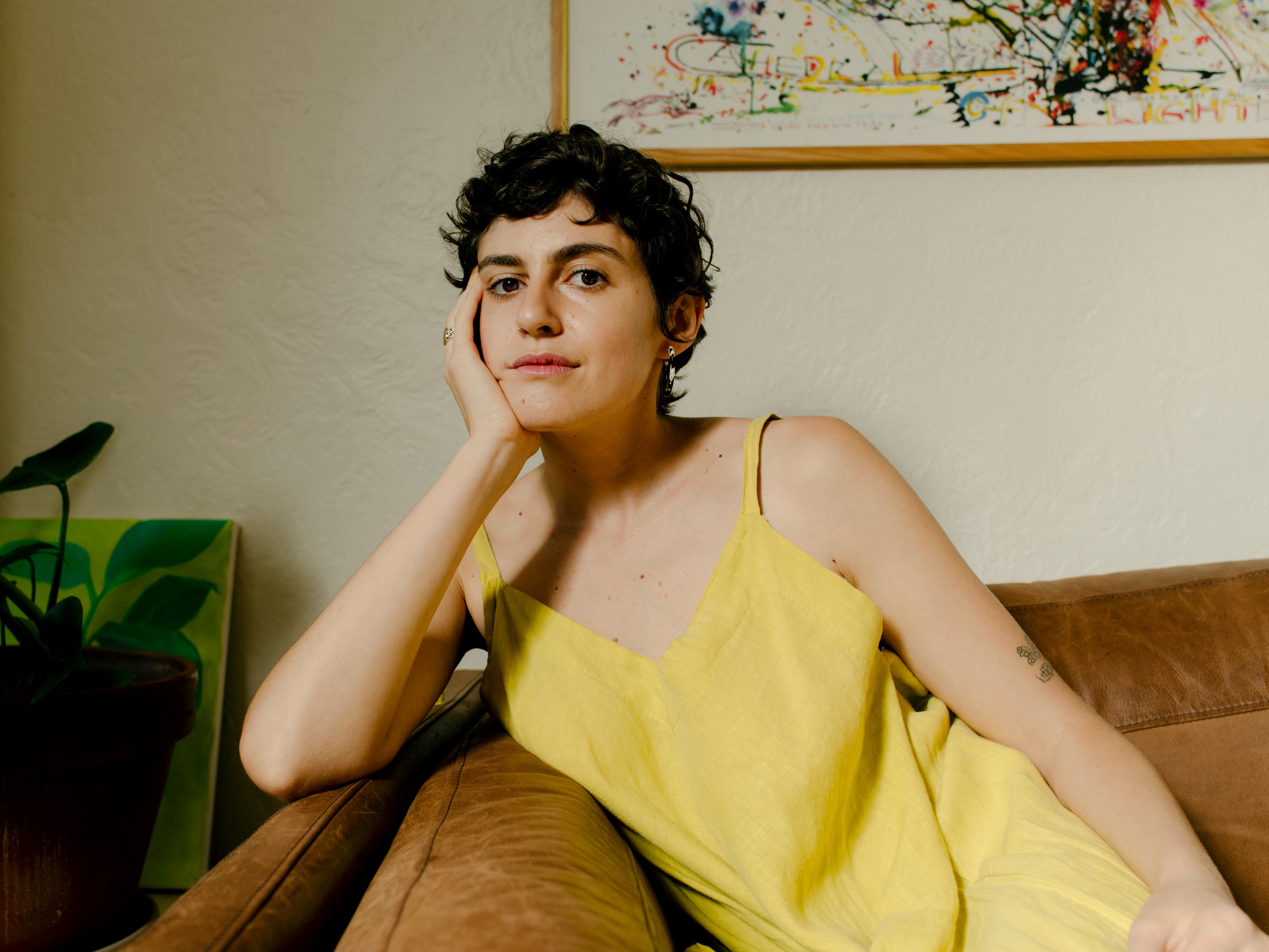 Zoe Schlanger wears a yellow dress and leans her head on her hand while sitting on a leather sofa next to a houseplant.