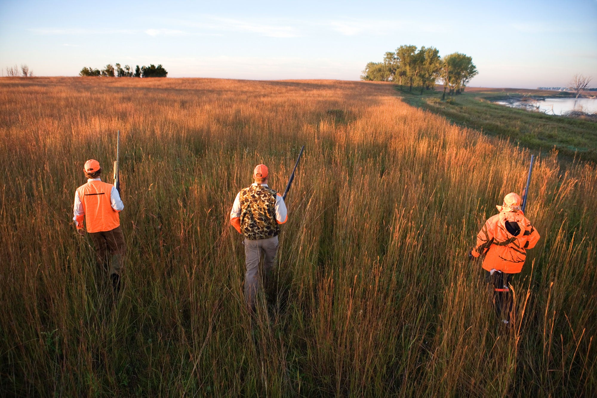 Three people in orange and camouflage walk away into a meadow while holding rifles.