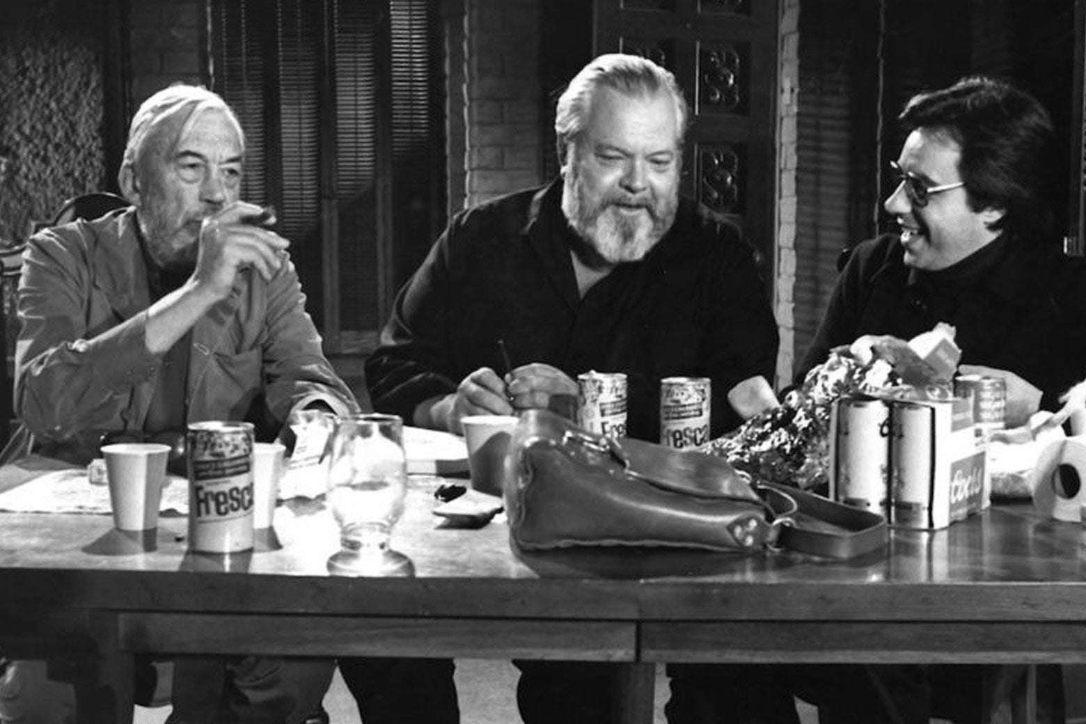 John Huston, Orson Welles, and Peter Bogdanovich in a scene from The Other Side of the Wind. They're seated at a table laden with cans of Fresca. Huston is smoking.