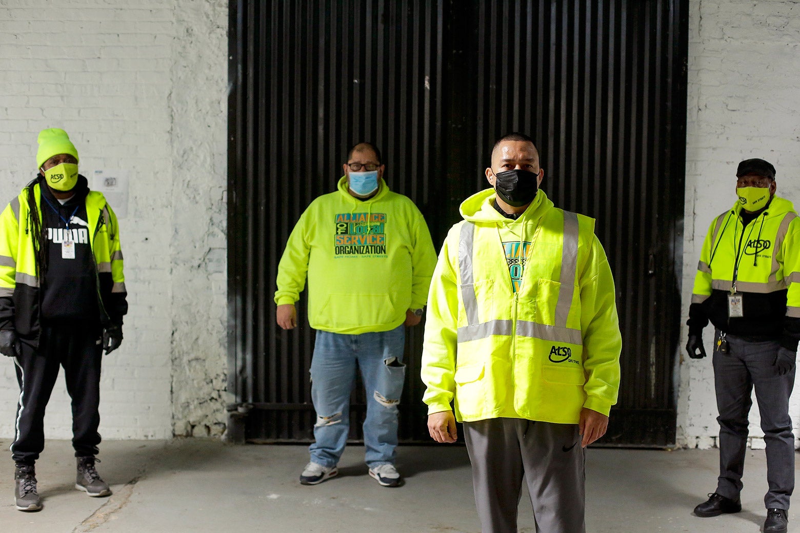 Four people wearing bright safety jackets and masks, standing a few feet apart from one another outside