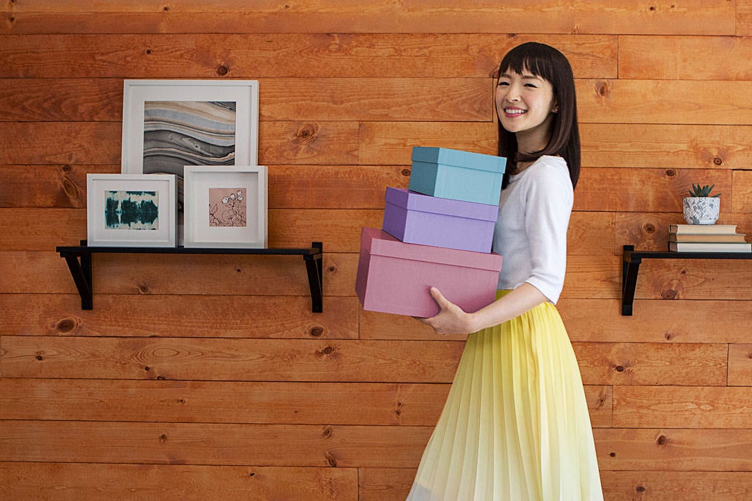 Marie Kondo smiling, carrying a neat stack of boxes, next to a wood wall with tasteful picture and plant mounts.