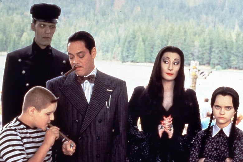 A tableau of the Addams family. Carel Struycken stands tall as the grey-skinned character Lurch in his butler uniform. Jimmy Workman, a child, is having a cigar in his striped t-shirt. Raul Julia, in his grey pinstripe suit, is lighting Workman's cigar, while smoking his own. Anjelica Huston looks over at them with widened eyes and her hands clasped together. Christina Ricci is frowning at the camera with her hair in two black braids.