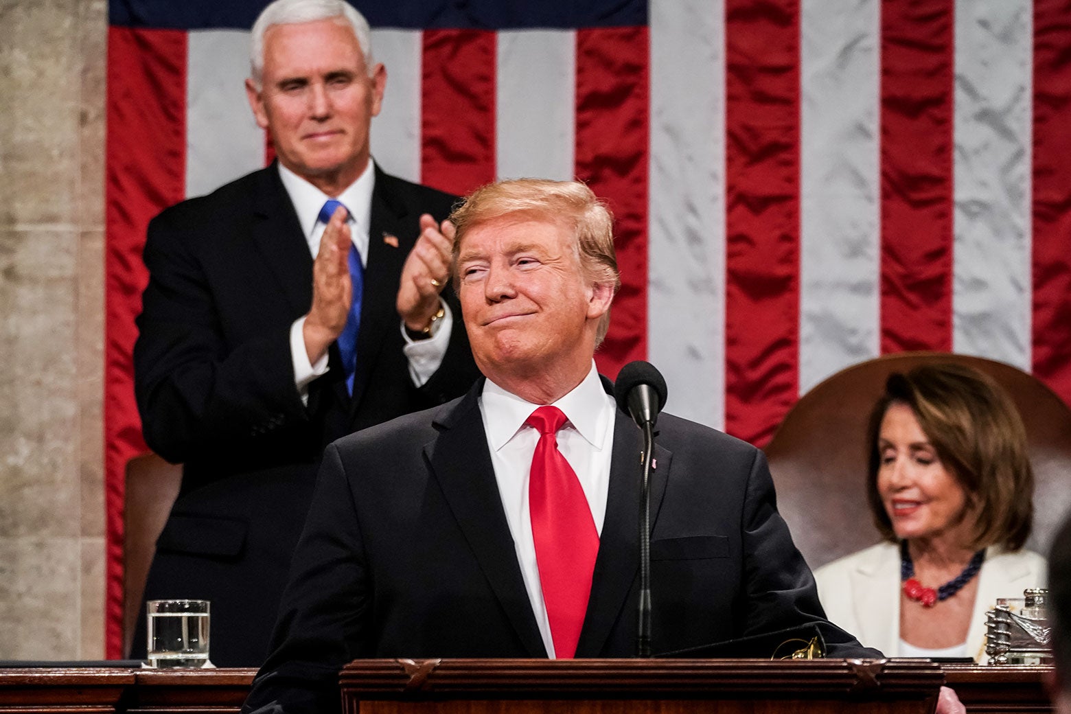 Donald Trump smiles as Mike Pence stands to applaud. Nancy Pelosi is seated and smiling.