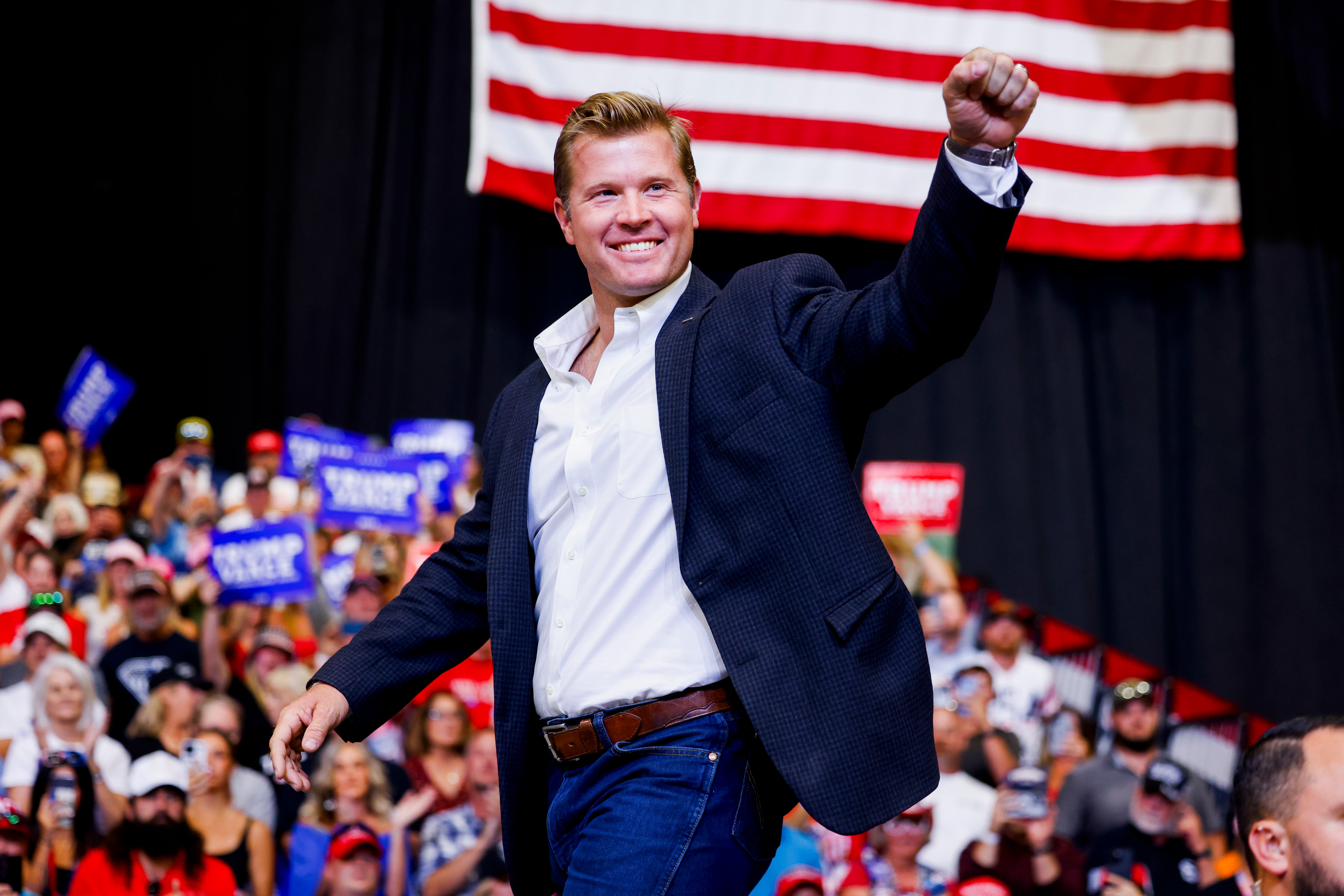 A white man in a navy suit smiles and raises his left hand in a fist onstage, with a U.S. flag and a crowd behind him.