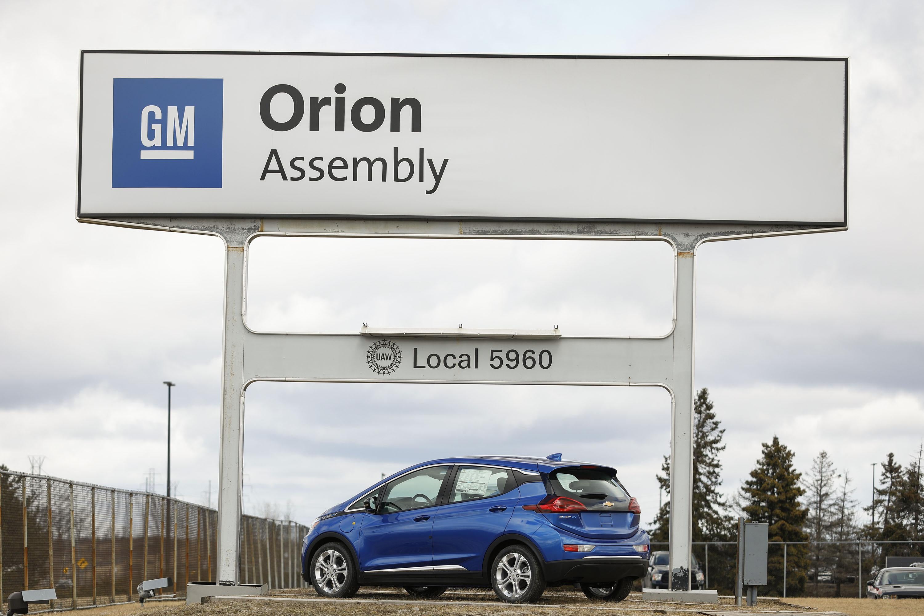 A blue electric vehicle is parked below the General Motors Orion Assembly plant sign.