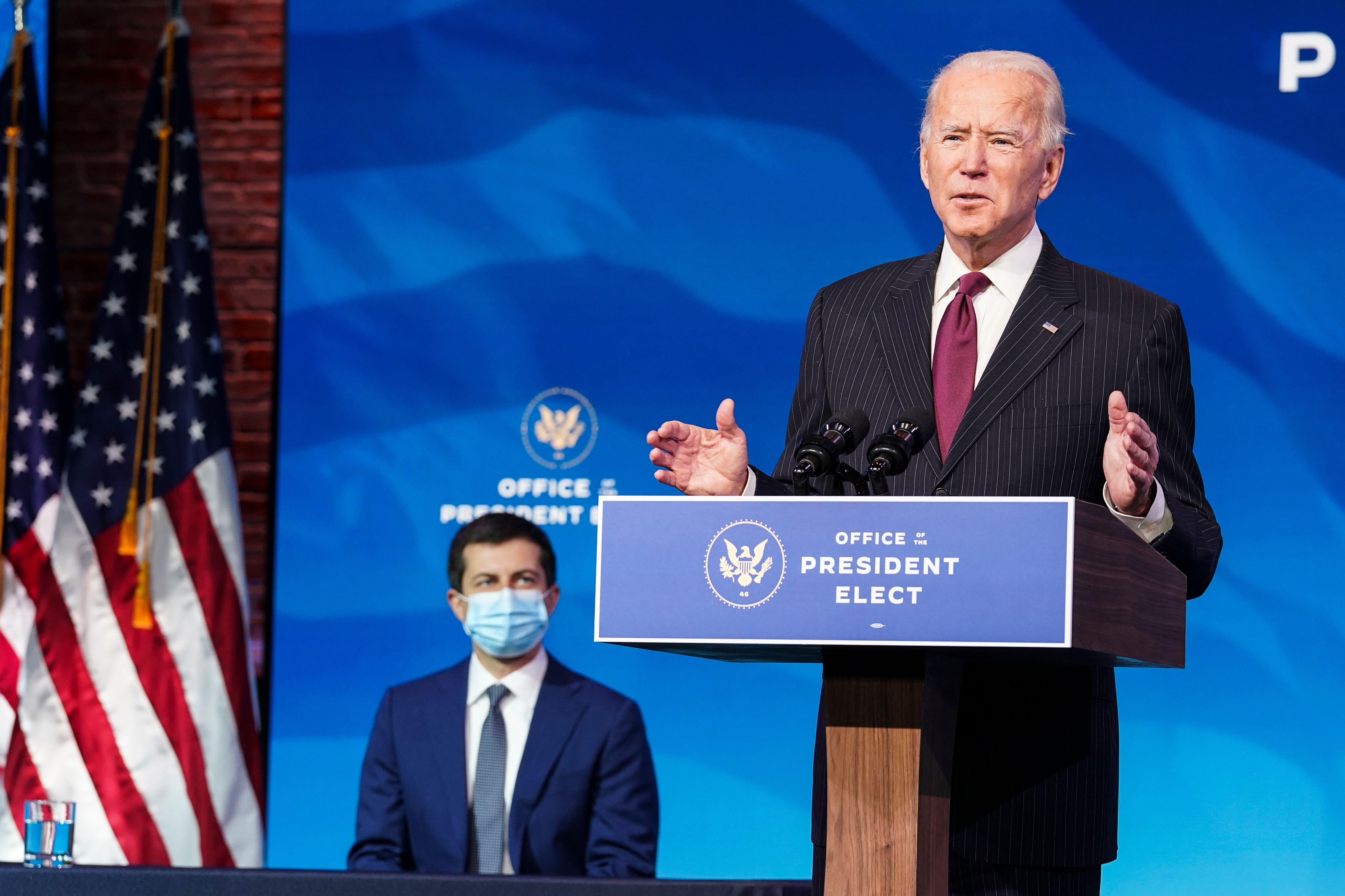 Biden speaks at a lectern while Buttigieg sits behind him, wearing a face mask.