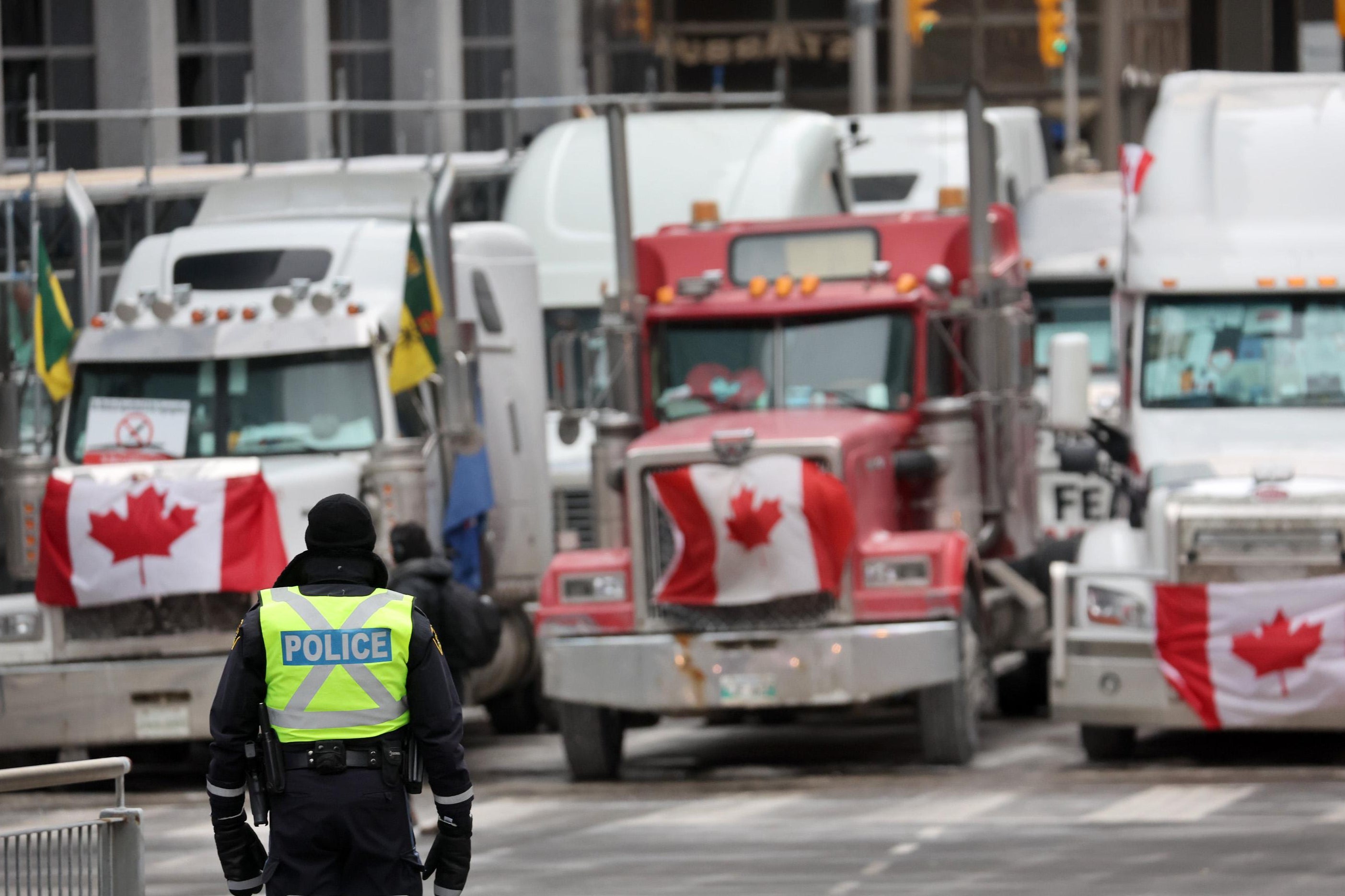 A police officer stands in front of three trucks with Canadian flags on their grilles that are parked in a downtown street