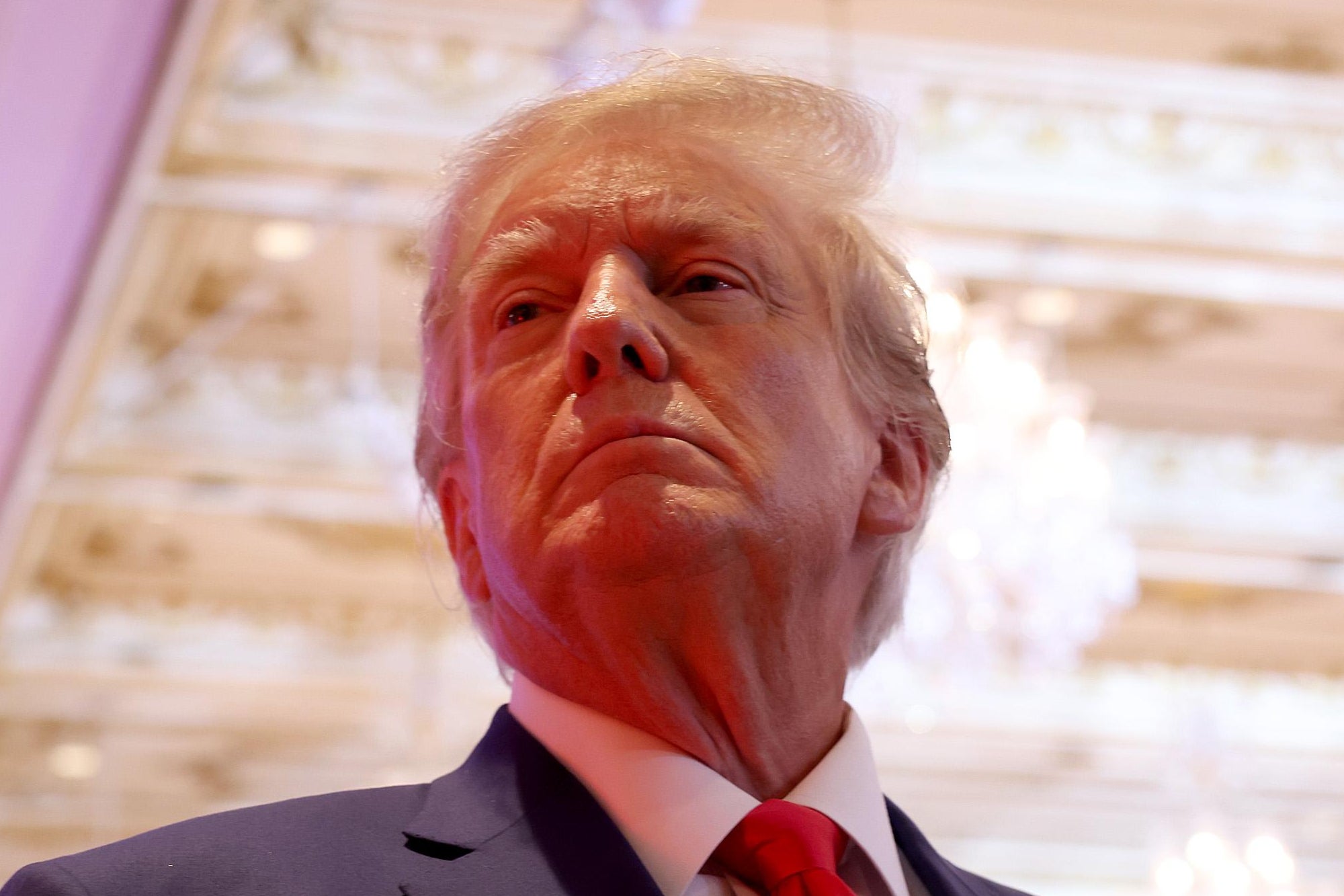 Trump looking upward in a suit, frowning close-mouthed, a fancy ceiling with chandelier behind him.