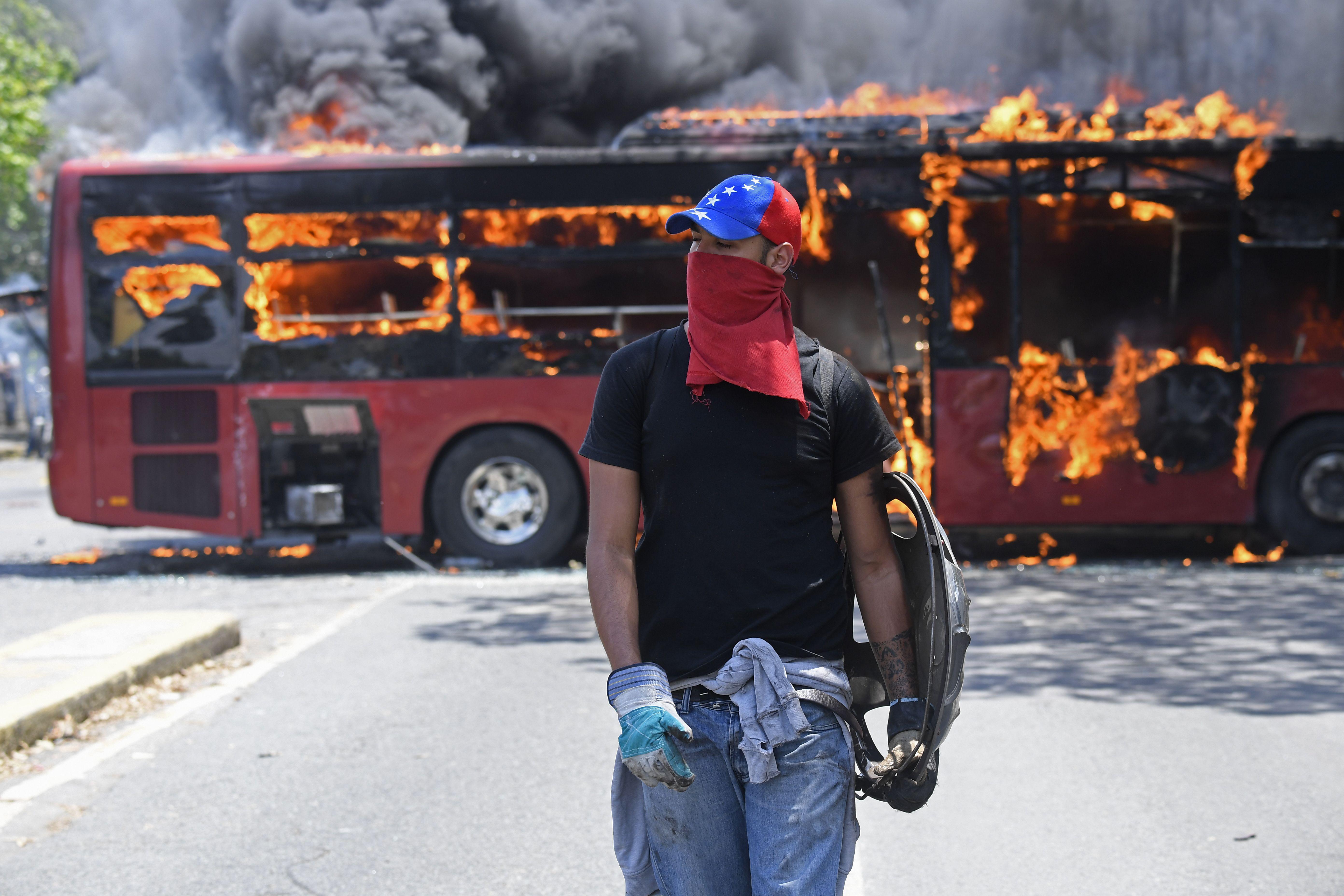 An opposition demonstrator wearing a hat and a cloth over his face walks in front of a burning bus.