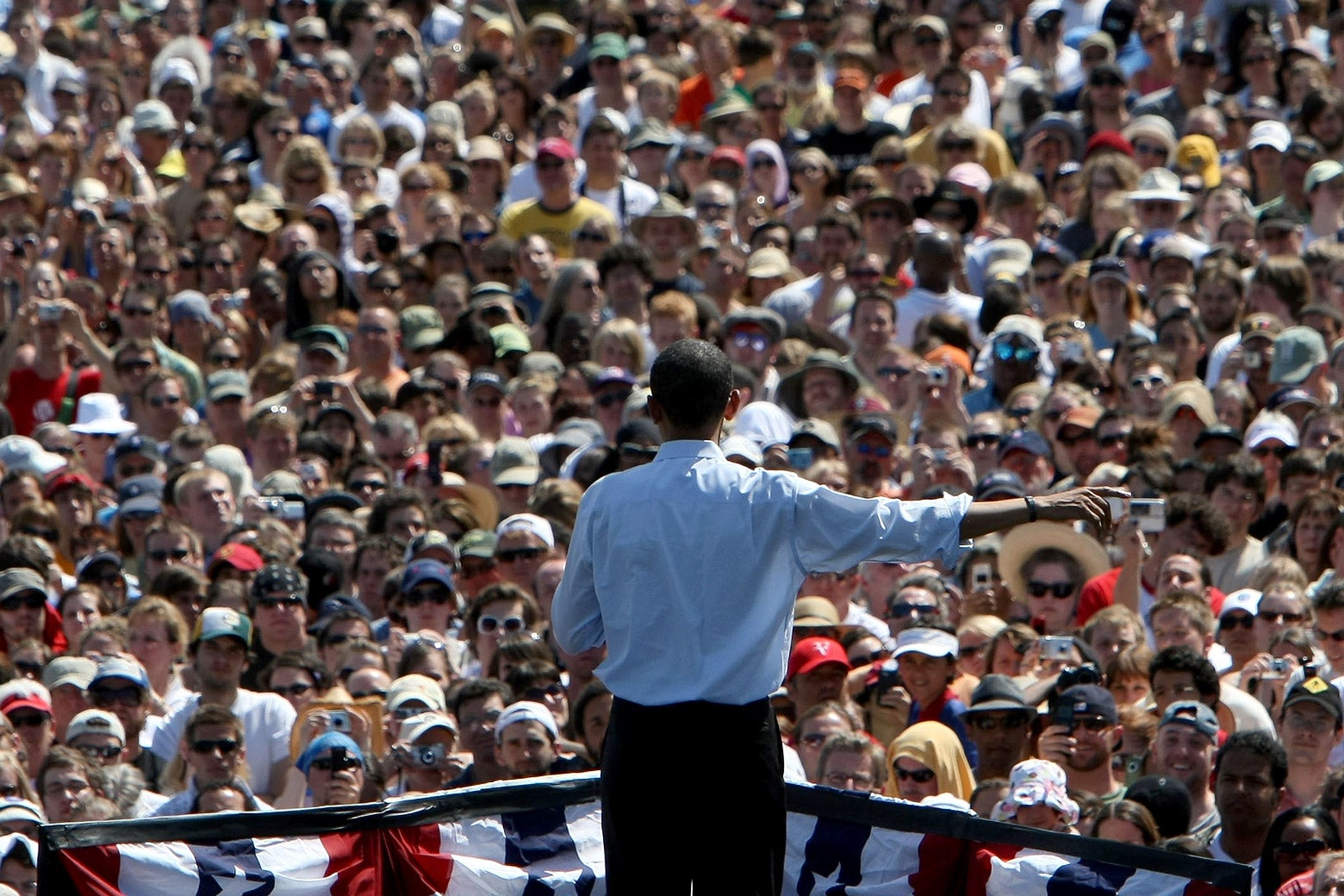 Barack Obama, seen from behind, gestures as he speaks to a large crowd outdoors.