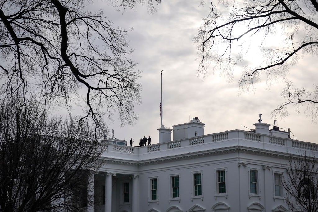 A flag at half-mast is seen above the upper floor of the White House against bare trees and a gray sky.