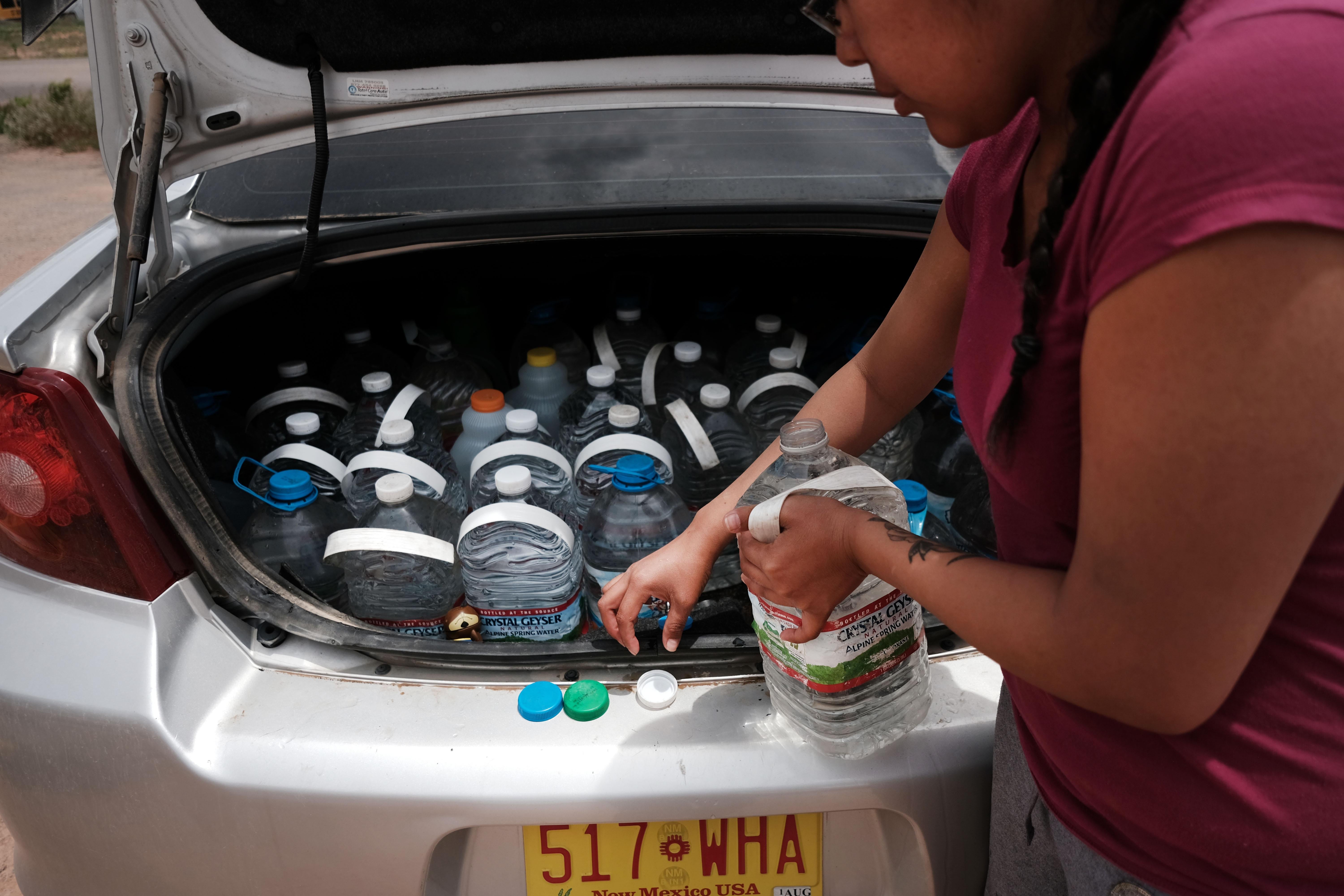A woman takes big bottle of water out of a car trunk full of plastic water bottles.
