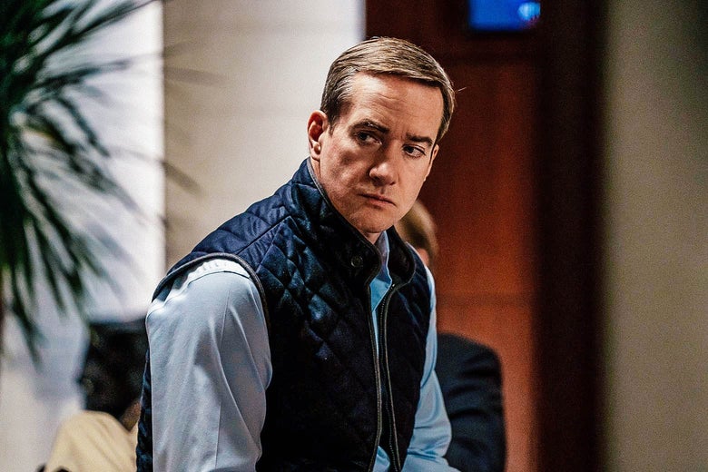 A man in a quilted vest looks toward his right while seated in some kind of office building.