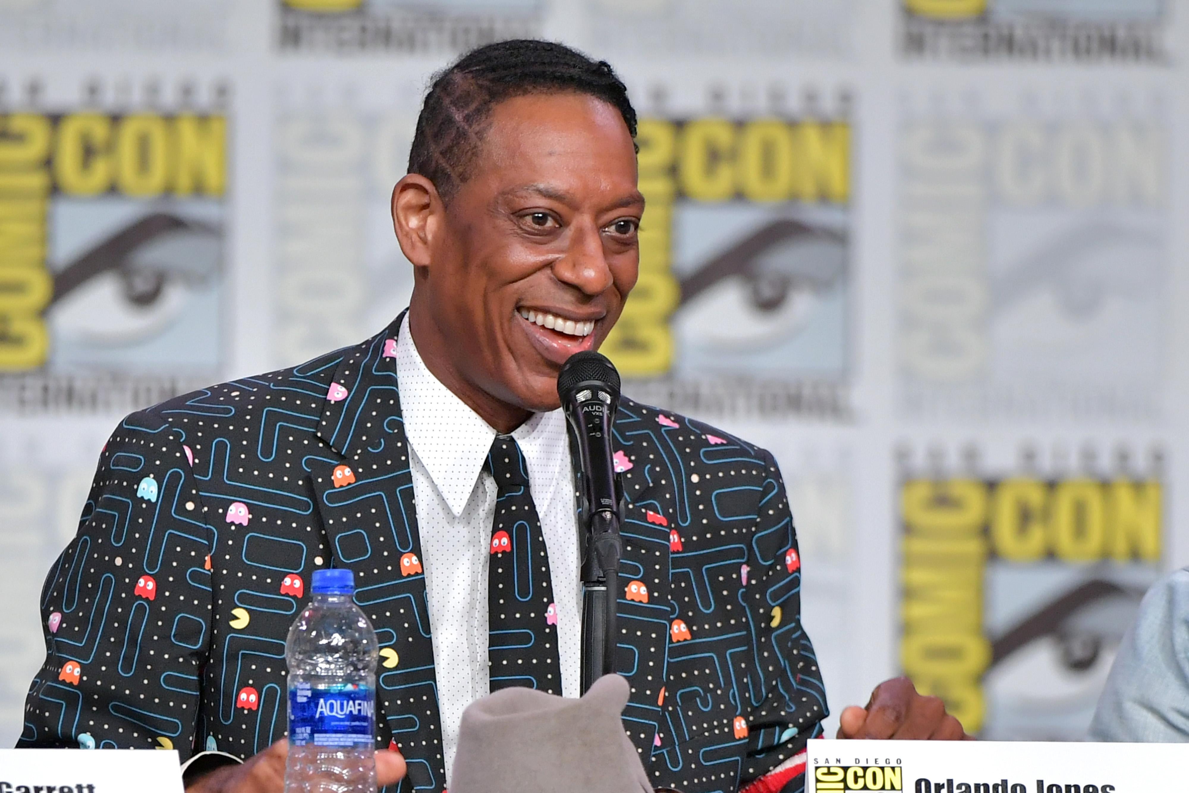 Orlando Jones, wearing a suit jacket with a Pac-Man maze on it, sits at a panel table before a Comic-Con logo backtrop, smiling broadly. 
