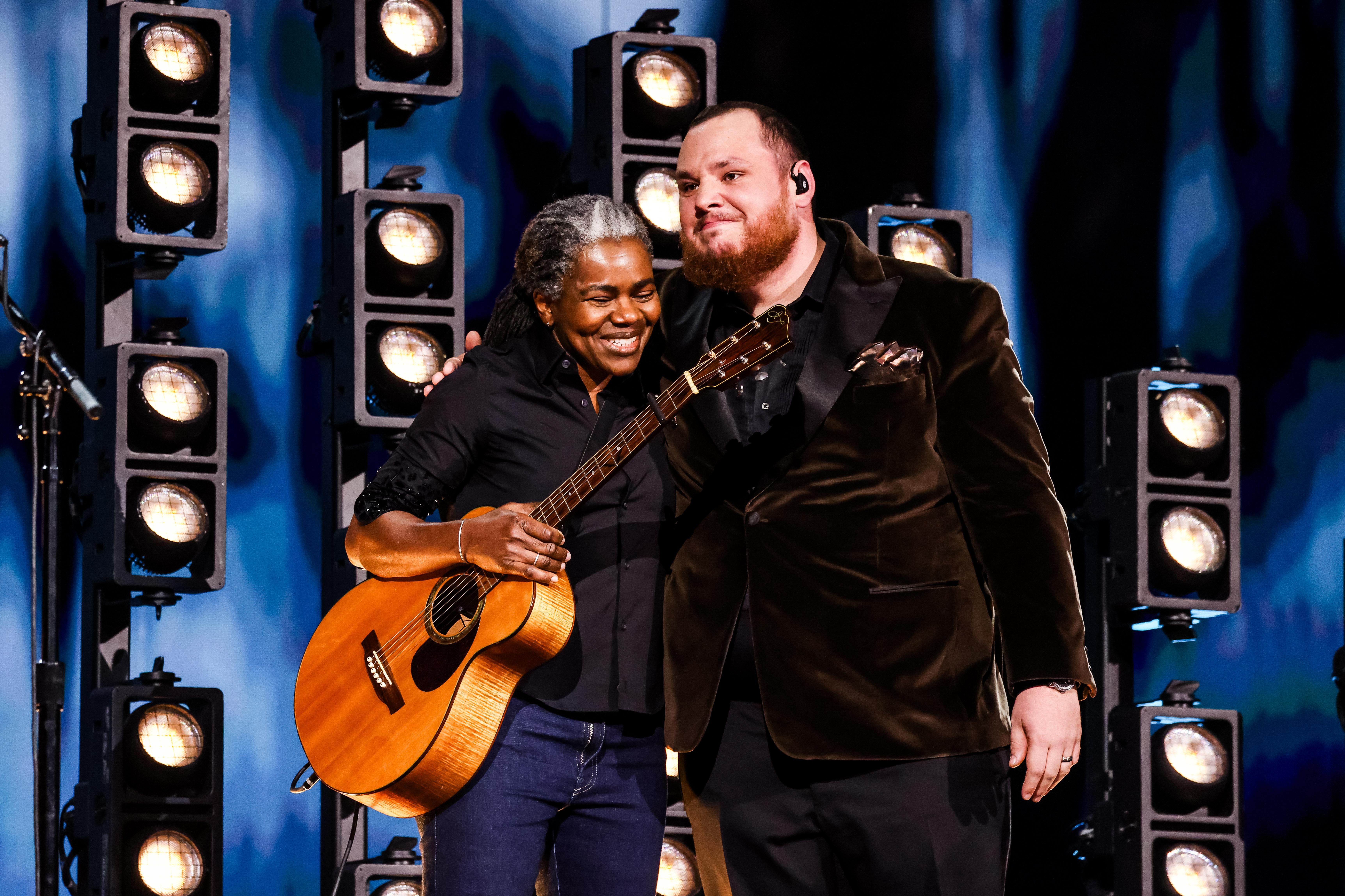 Tracy Chapman, holding a guitar, smiles and places her arm around Luke Combs, who looks on.