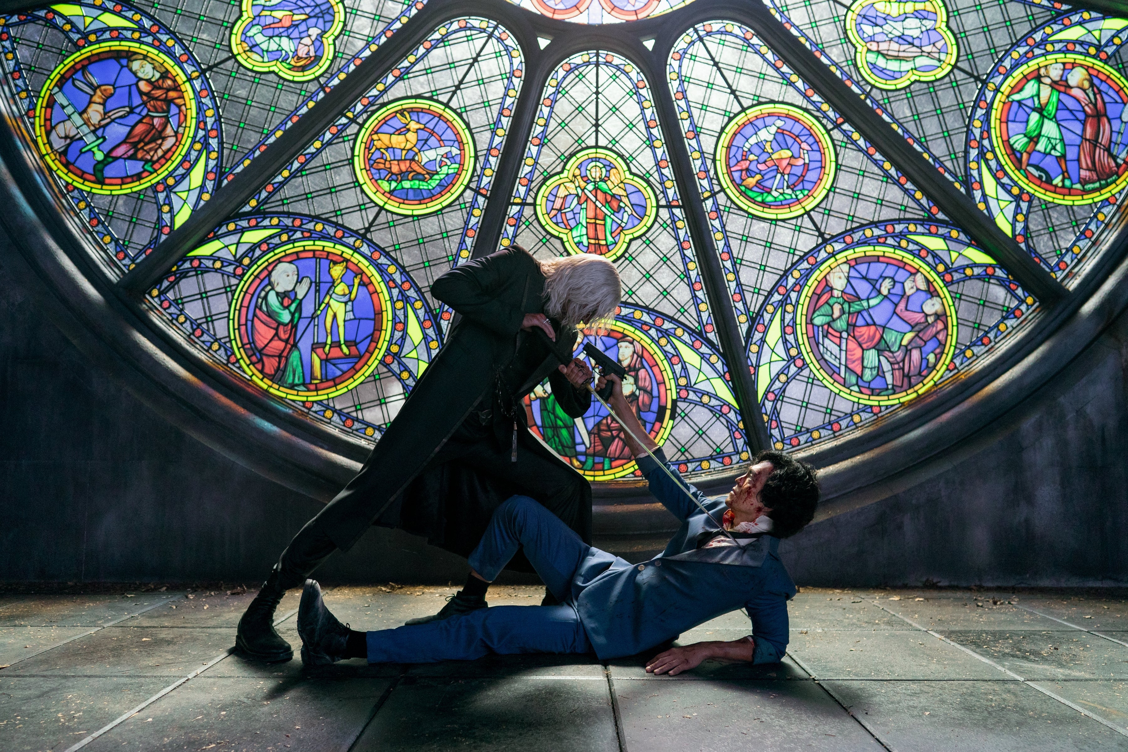 One man hunches over and holds a sword to the neck of a man lying on the ground who is pointing a gun up at him. Behind them is a huge stained glass window.