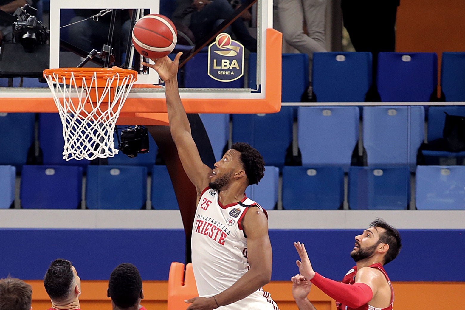 Mitchell lays the ball into the hoop (or grabs a rebound) as other players look on below him. The stadium seats behind the hoop are empty.