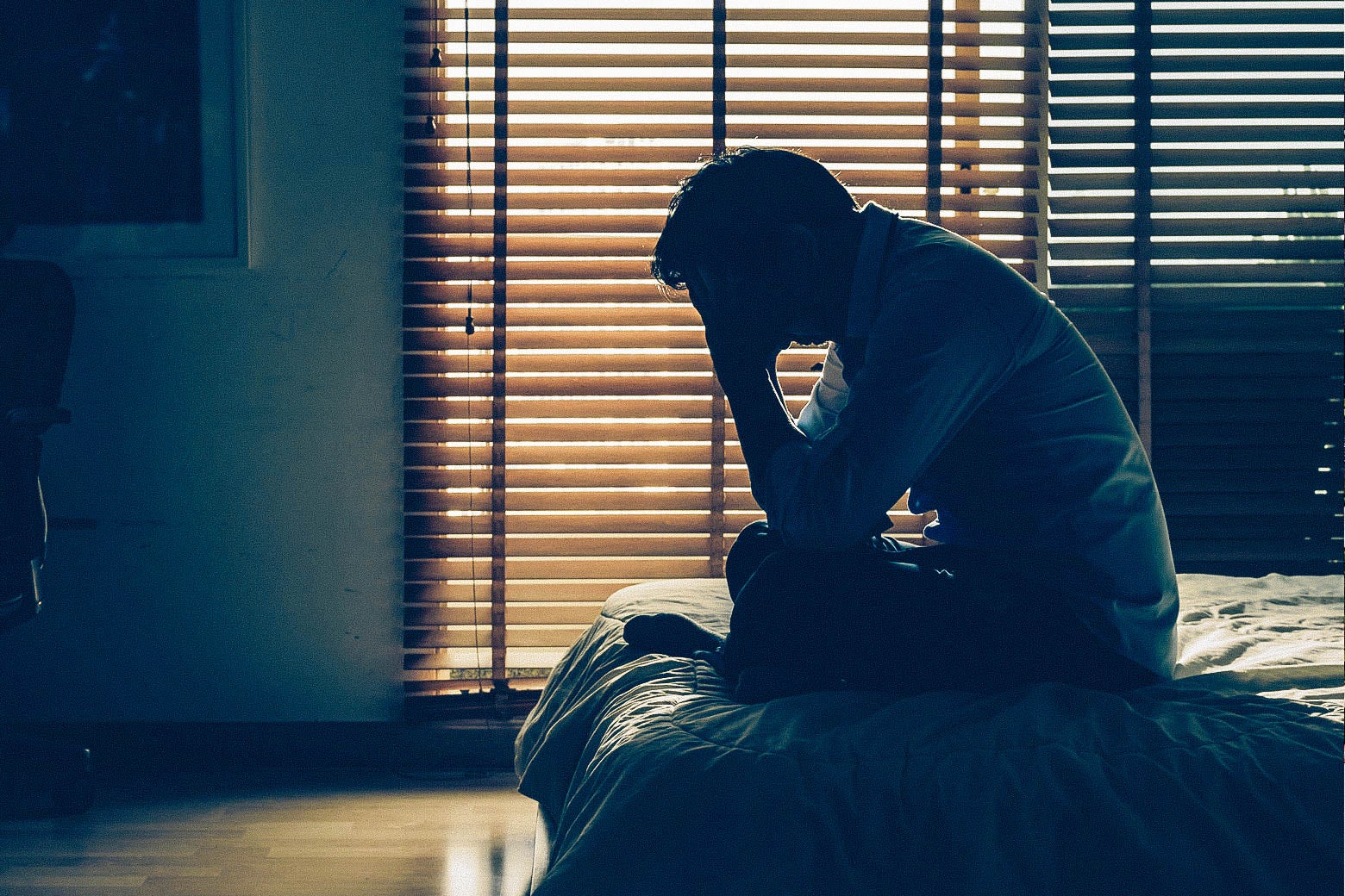 Stock image of a man sitting on a bed, holding his head with one hand despondently.