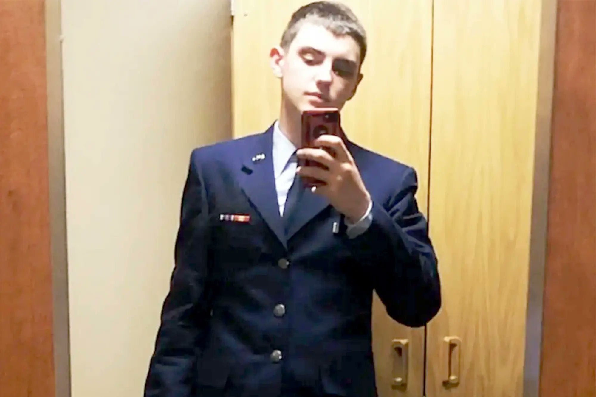 A young man with light skin and brown hair, wearing a navy blue airman's uniform, takes a mirror selfie with a cell phone.