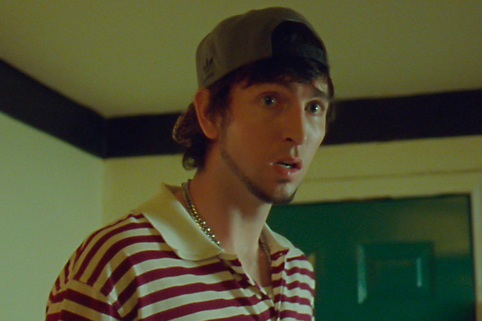 Nicholas Braun in a backward baseball cap and striped polo with a thin chin-strap beard.