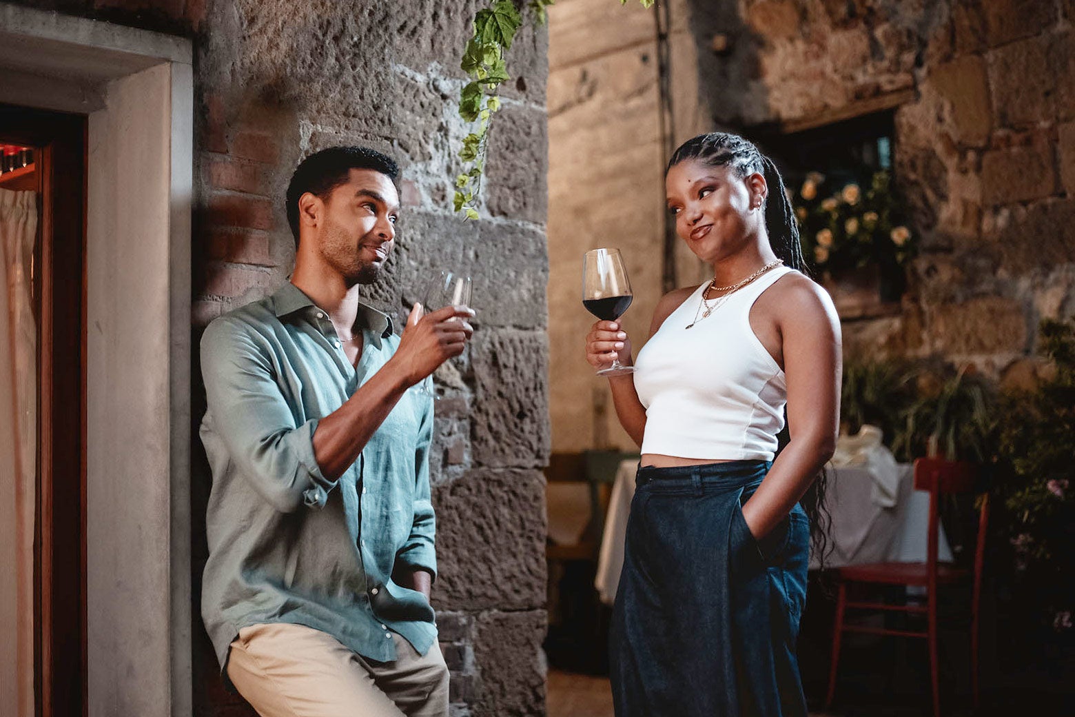 Regé-Jean Page and Halle Bailey in You, Me & Tuscany, holding glasses of wine in an Italian village setting.