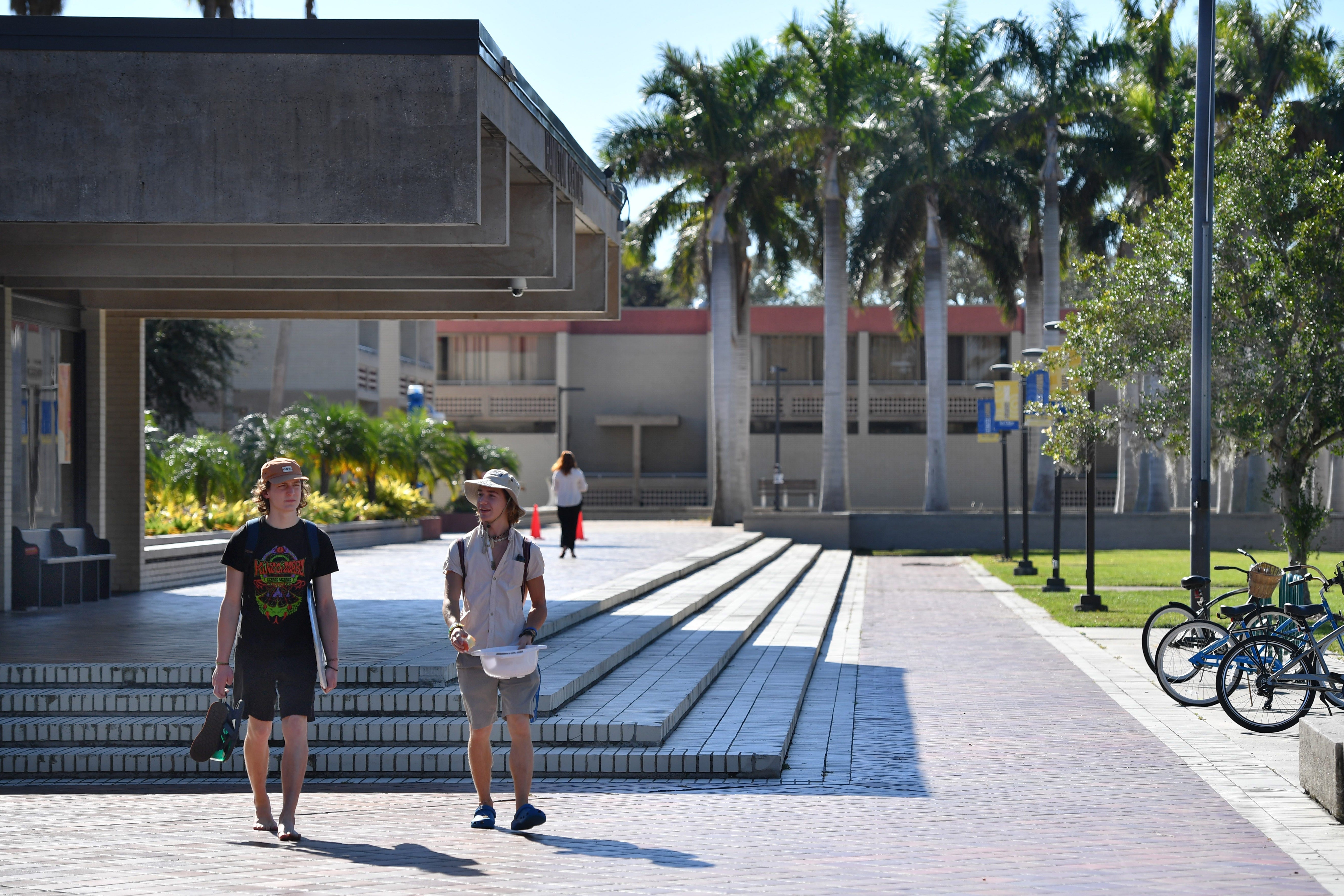 Two students in shorts walk near a brutalist-style building on a sunny day. A bike rack, another building, and palm trees are seen in the background.