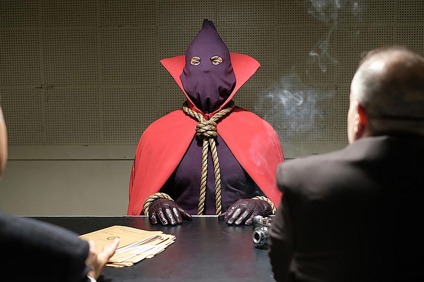 Hooded Justice sits at a table during a police interrogation.