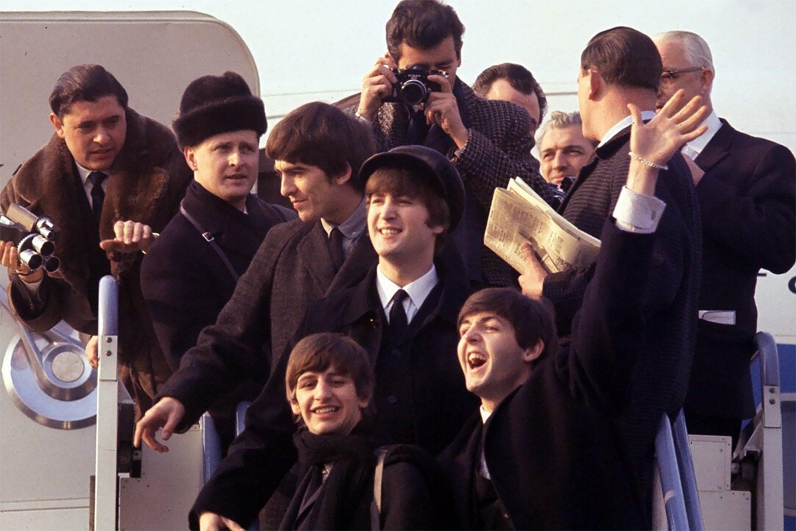 A shot of the Beatles waving from airplane steps as photographers take pictures. 