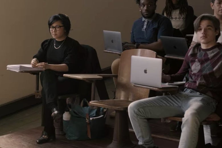 A teaching assistant and students sit in wooden seats in a college classroom.