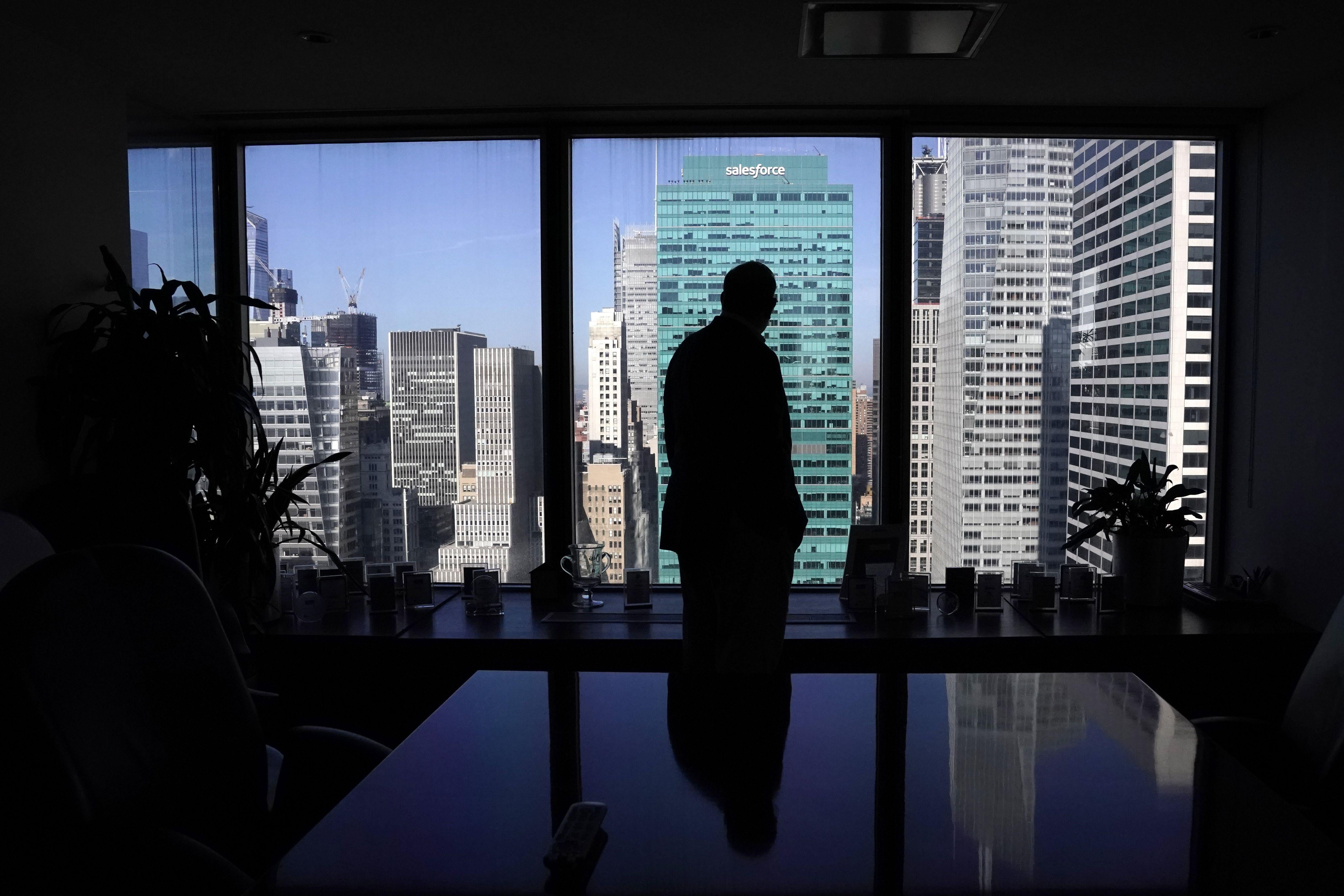 A man in a suit stands backlit in front of a window in an empty office overlooking Fifth Avenue buildings. 