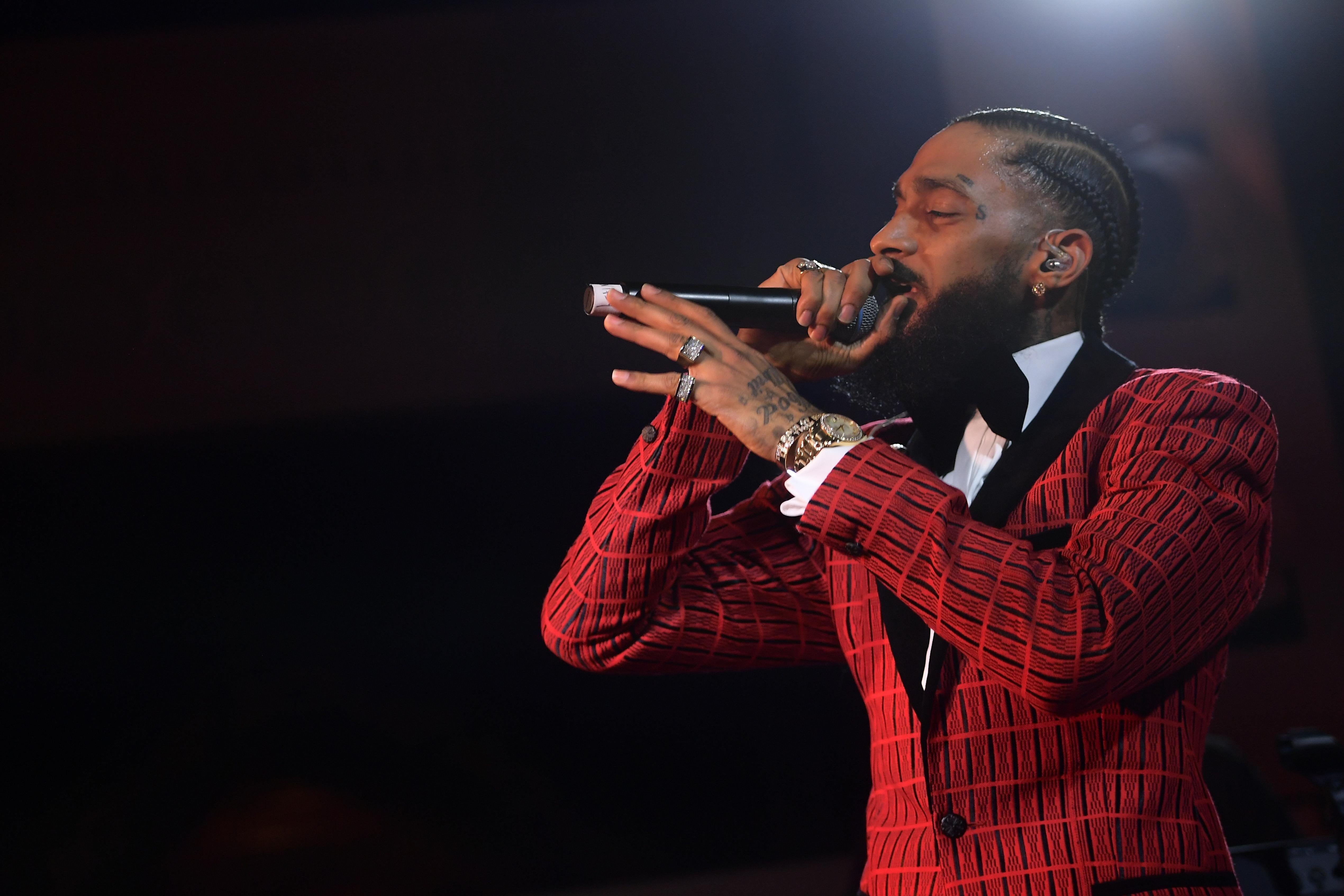 Nipsey Hussle, wearing a red jacket and black bow tie, holding a mic to his mouth as he performs. 