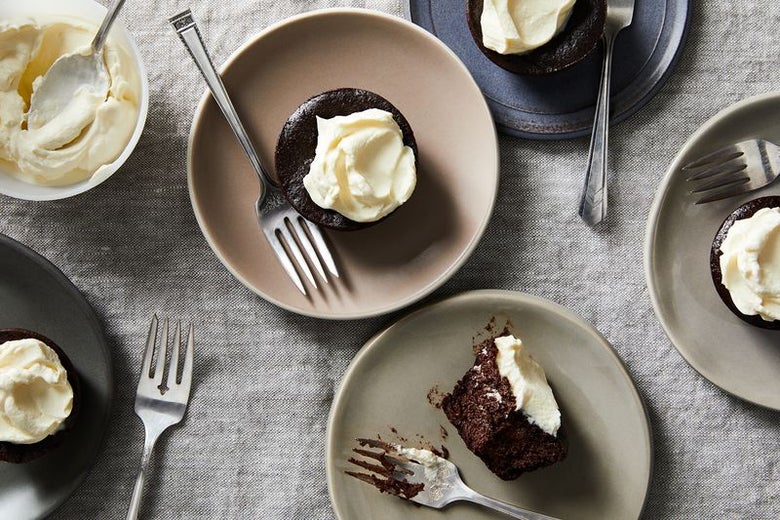 Several dark brown chocolate cup cakes with luscious white frosting sit on individual plates with forks next to a bowl of the frosting