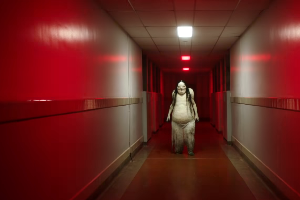 A monsterous figure standing in a dim red lit hallway, in a still from the film adaptation of Scary Stories.