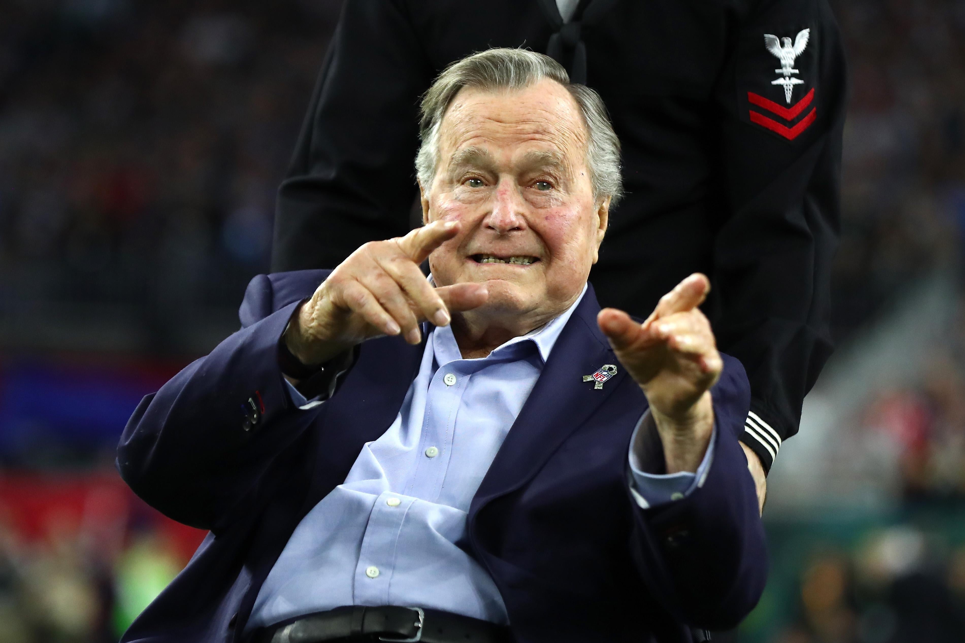 George H W Bush points to the crowd. Someone wearing a military uniform wheels his chair behind him. 