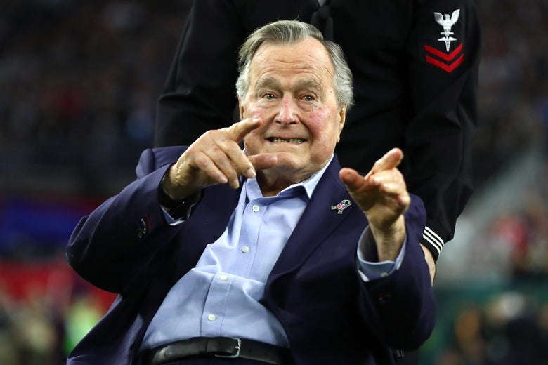 George H W Bush points to the crowd. Someone wearing a military uniform wheels his chair behind him. 
