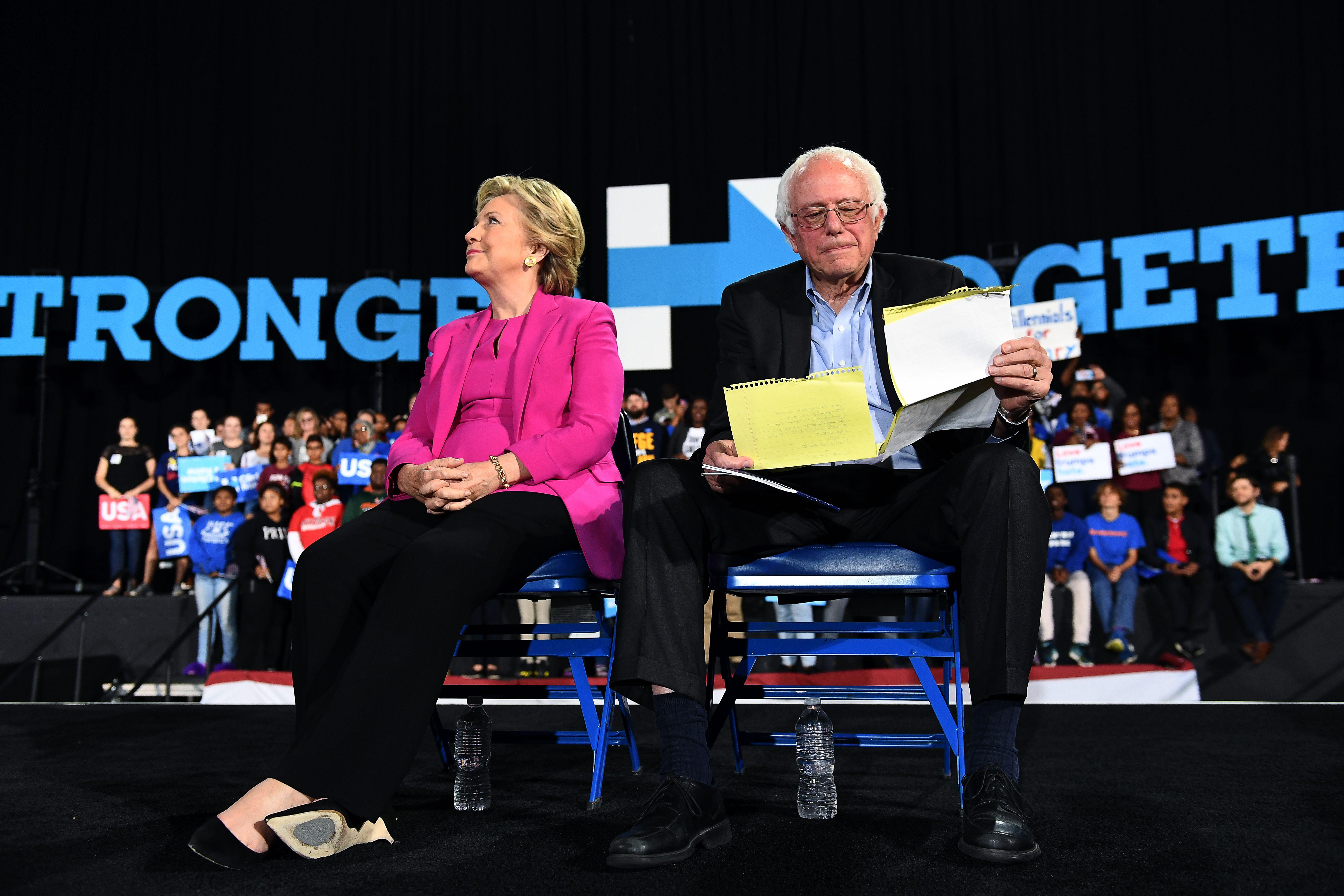 Hillary Clinton and Bernie Sanders sit on chairs looking away from one another during a campaign rally. Bernie is looking at sheets of notes.