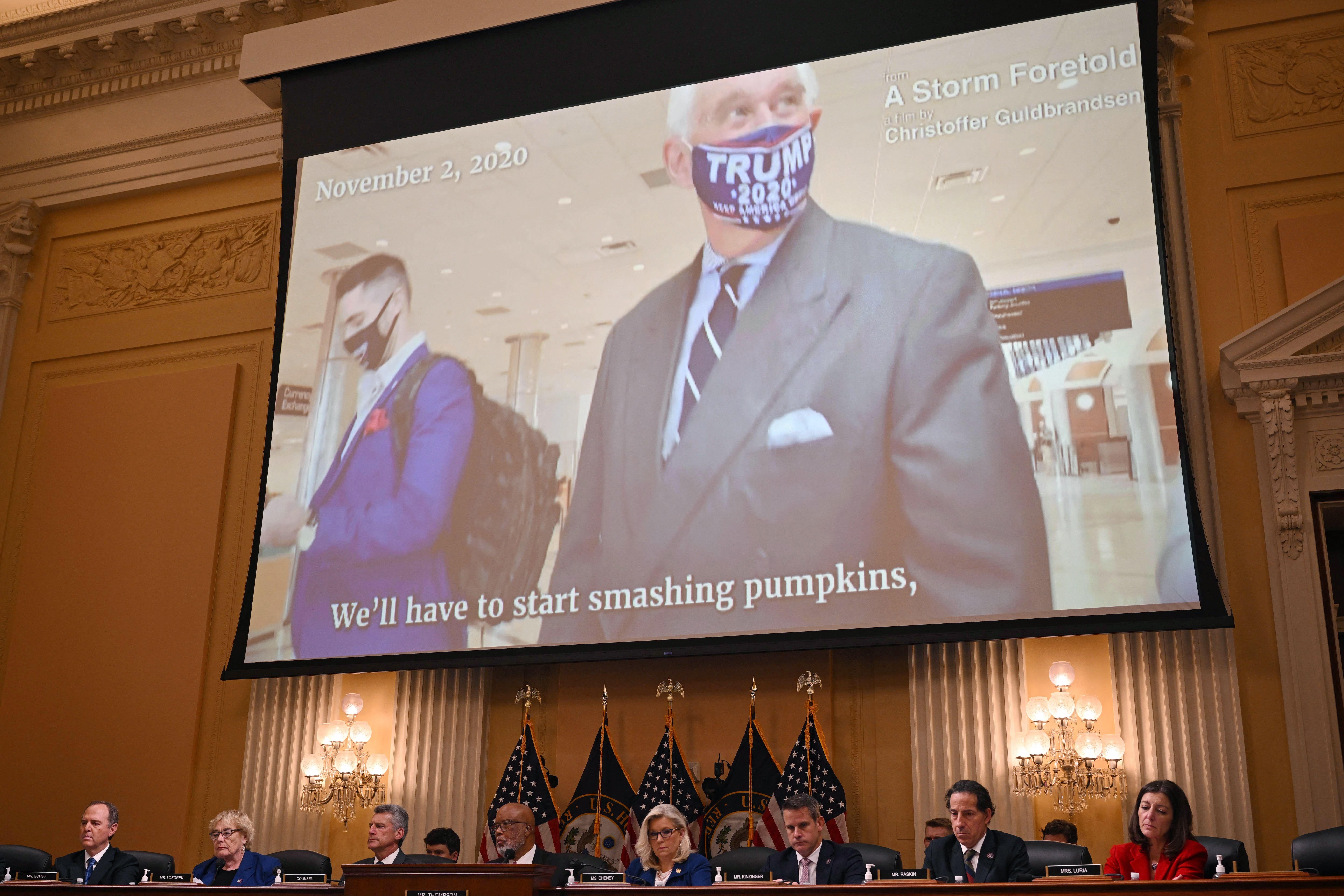 Roger Stone in a Trump 2020 mask on a big screen behind the House committee on Jan. 6, at their Thursday hearing. The closed captioning on the screen reads Stone's quote "We'll have to start smashing pumpkins."
