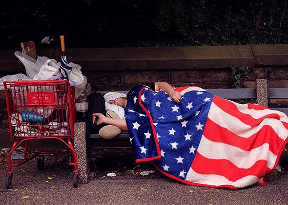 A homeless man sleeps under an American Flag blanket.