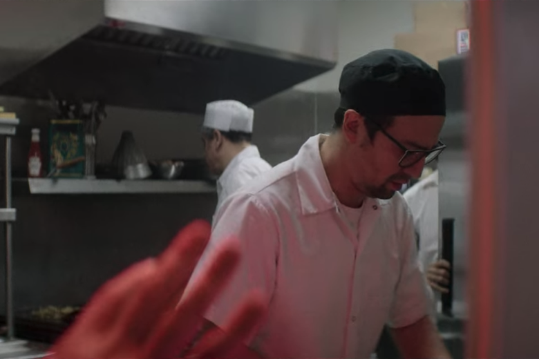 Lin-Manuel Miranda, can be seen through a diner kitchen window dressed as a line cook looking down.