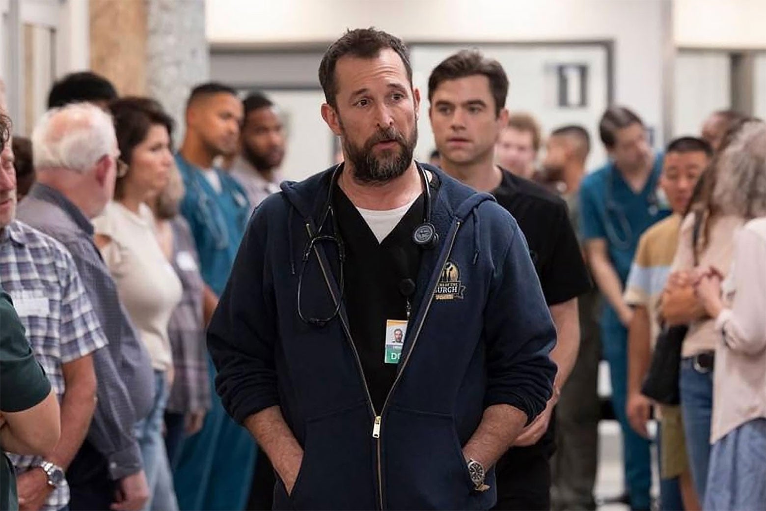 A doctor in a blue sweatshirt and wearing a stethoscope walks through the hallway of a crowded ER with his hands in his pockets, looking concerned.