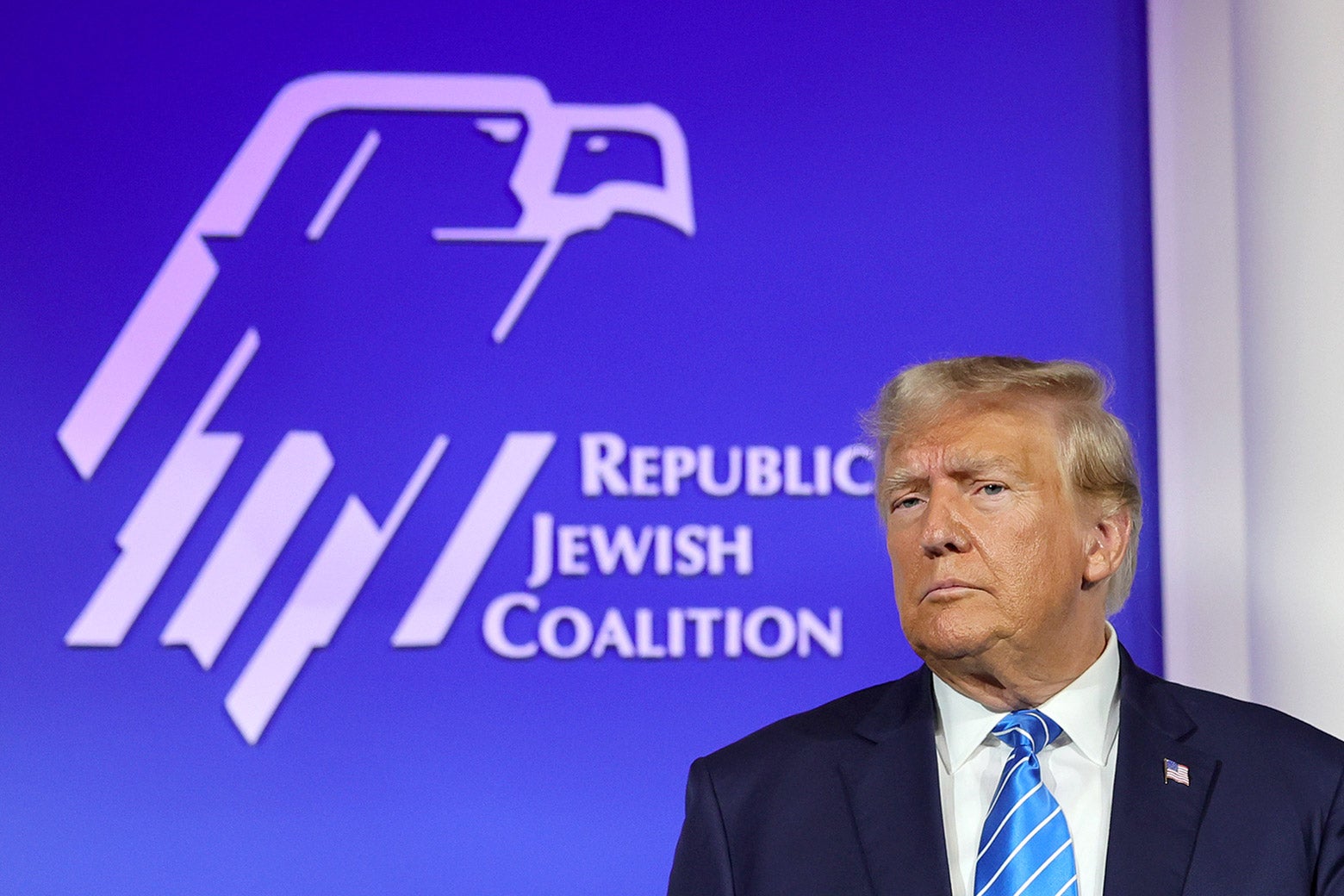 Trump furrows his brow and frowns in front of the logo for the Republican Jewish Coalition, an eagle with a Star of David embedded in it.